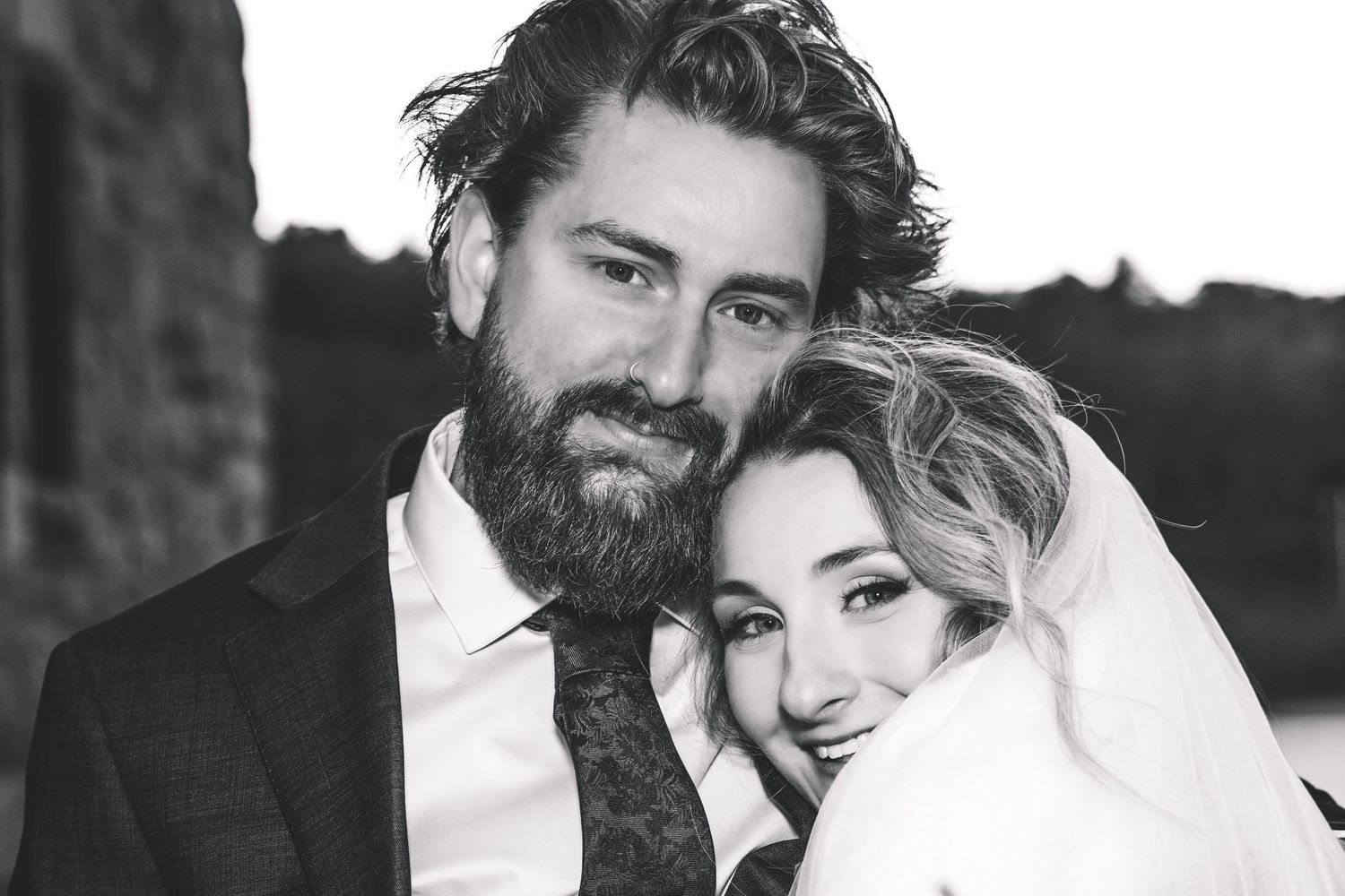 Black and white wedding portrait of a bearded groom in a suit and bride in a veil embracing outdoors.