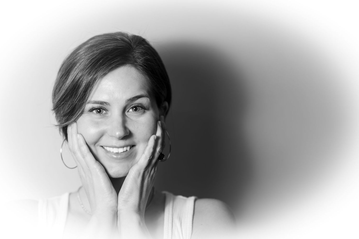Black and white portrait of a smiling young woman with hands on her cheeks against a light background.