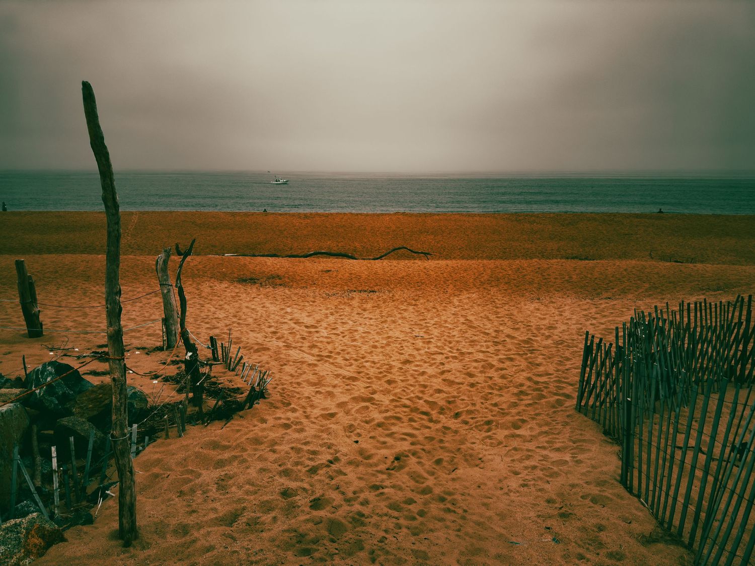 A sandy beach path lined with wooden fences leads to a calm ocean under an overcast sky.