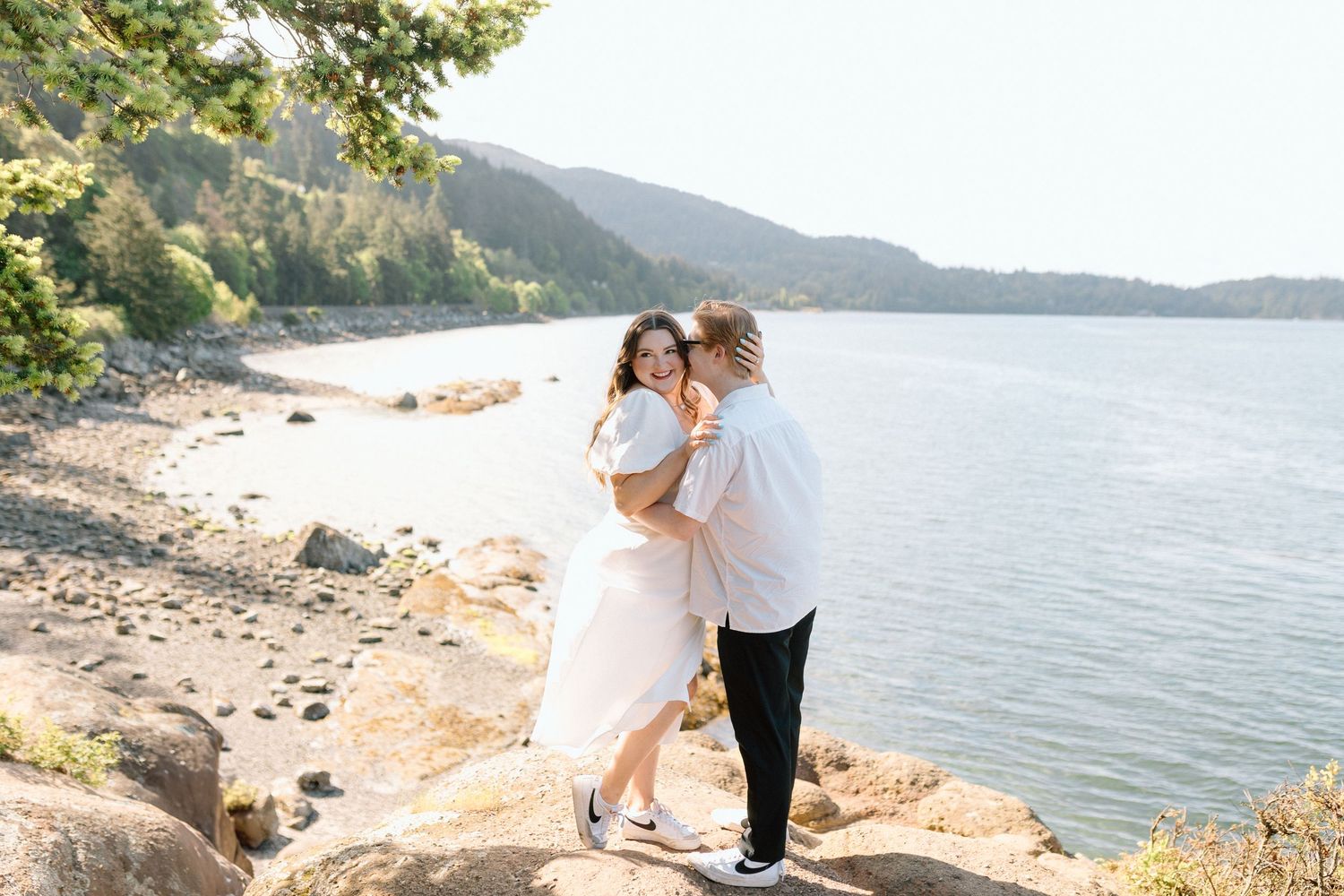 Romantic couple embraces on rocky coastline with mountain and ocean views at sunset.