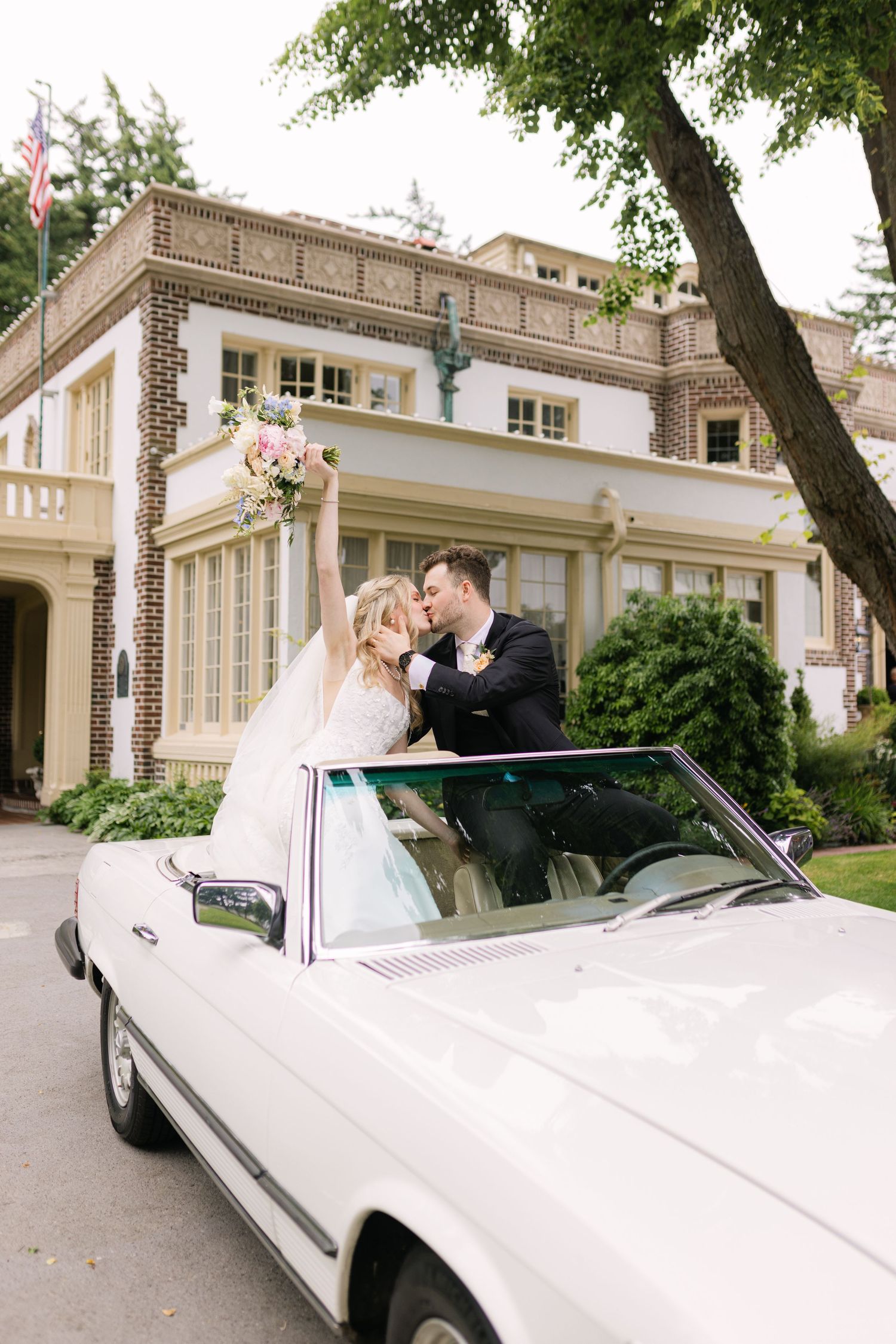 Classic white convertible parked in front of an elegant mansion with stone architecture and manicured grounds.
