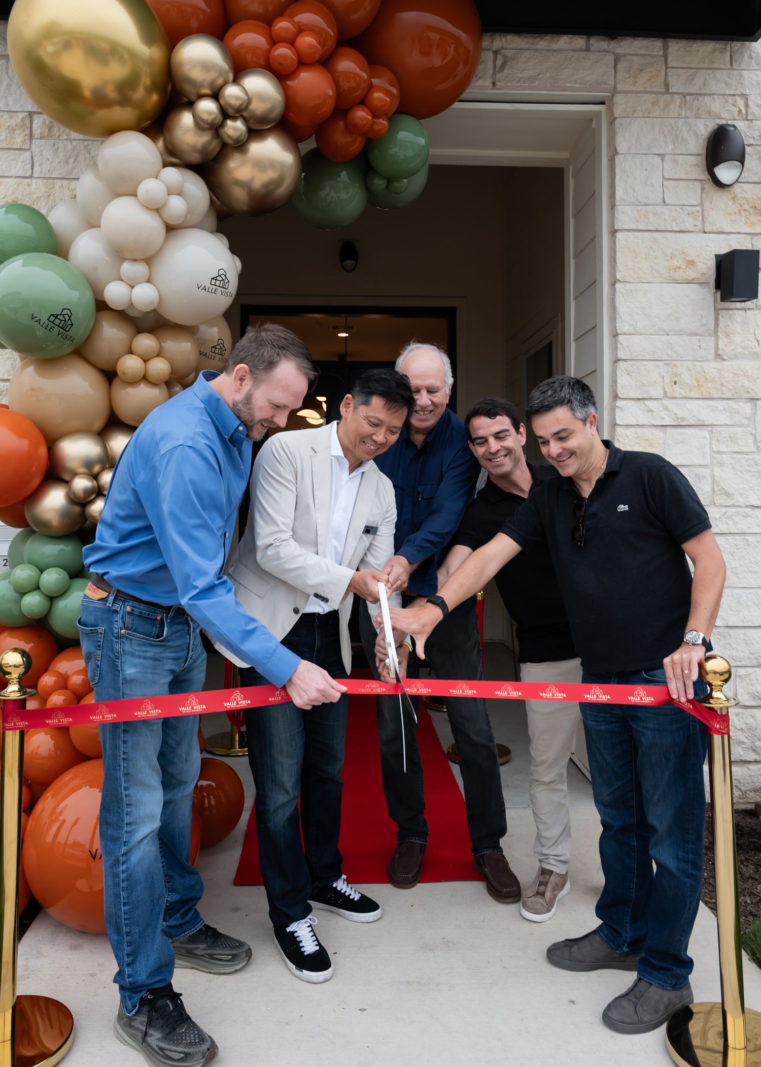 Five men cutting a red ribbon at a grand opening ceremony with colorful balloon arch decorations.