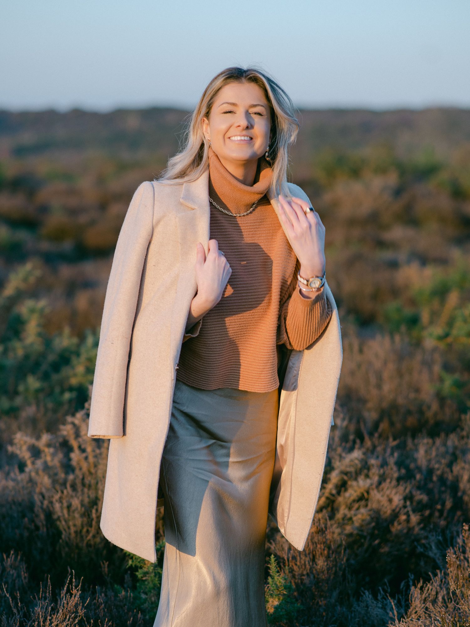 Smiling blonde woman in beige coat and brown turtleneck sweater standing in golden hour sunlight outdoors.
