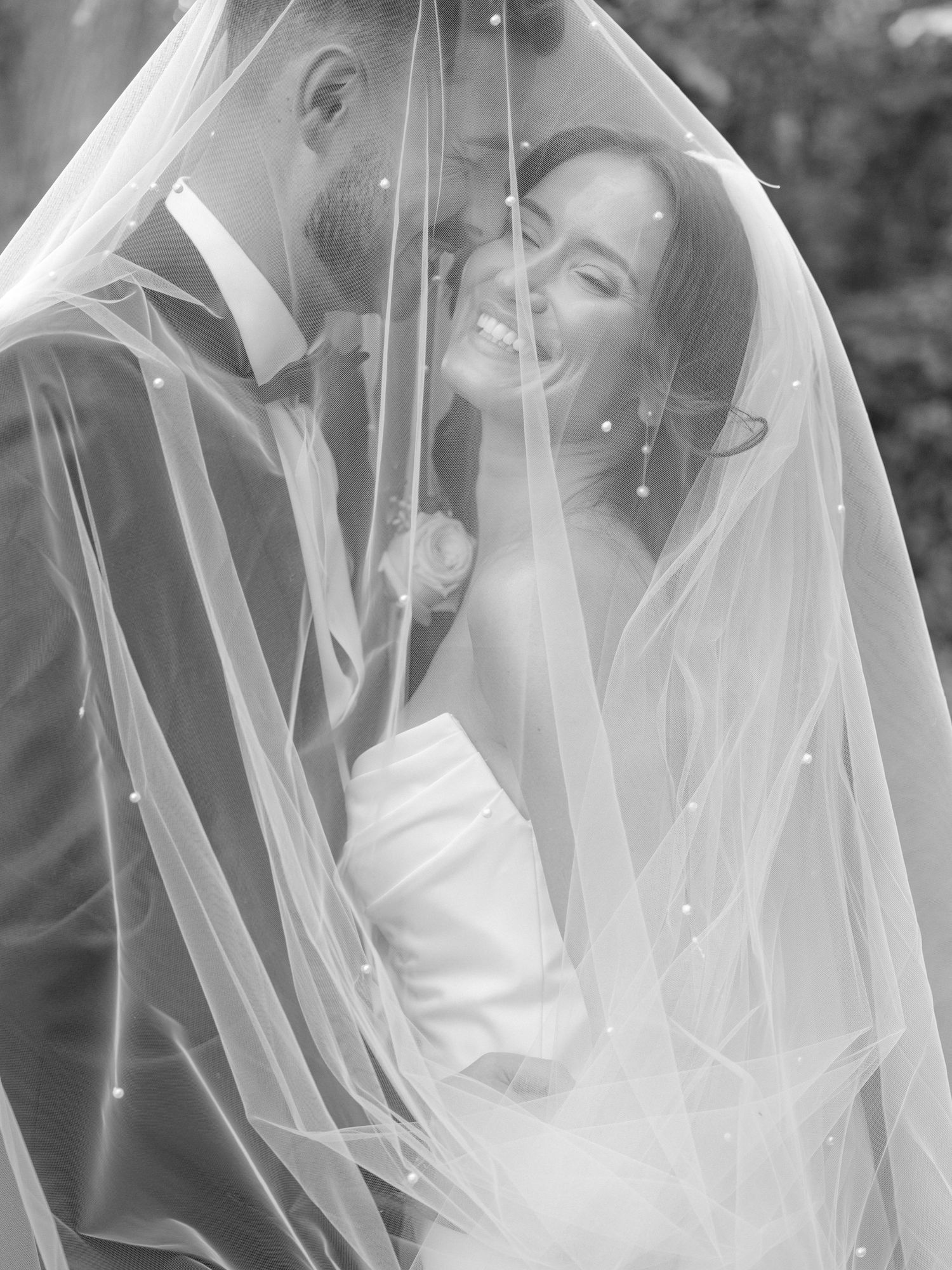Black and white wedding photo of smiling bride under veil with groom, romantic couple portrait.