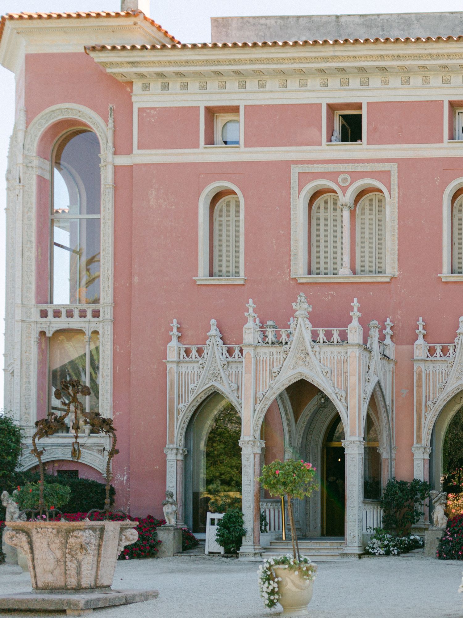 Pink historic villa with ornate Gothic arches, cream stone details, and arched windows in a Mediterranean courtyard setting.