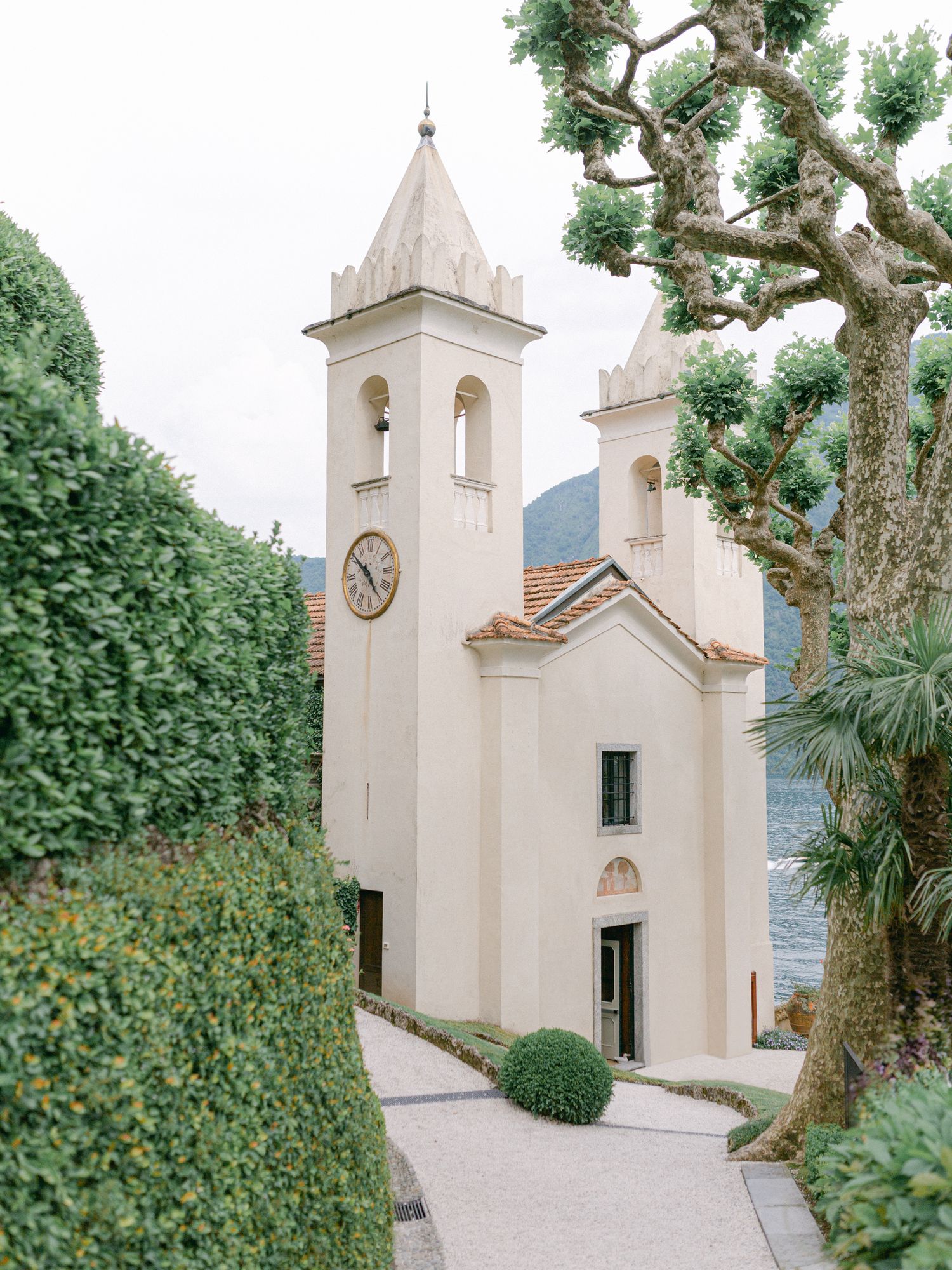 A charming Italian chapel with a clock tower, surrounded by manicured hedges and trees, overlooking Lake Como.