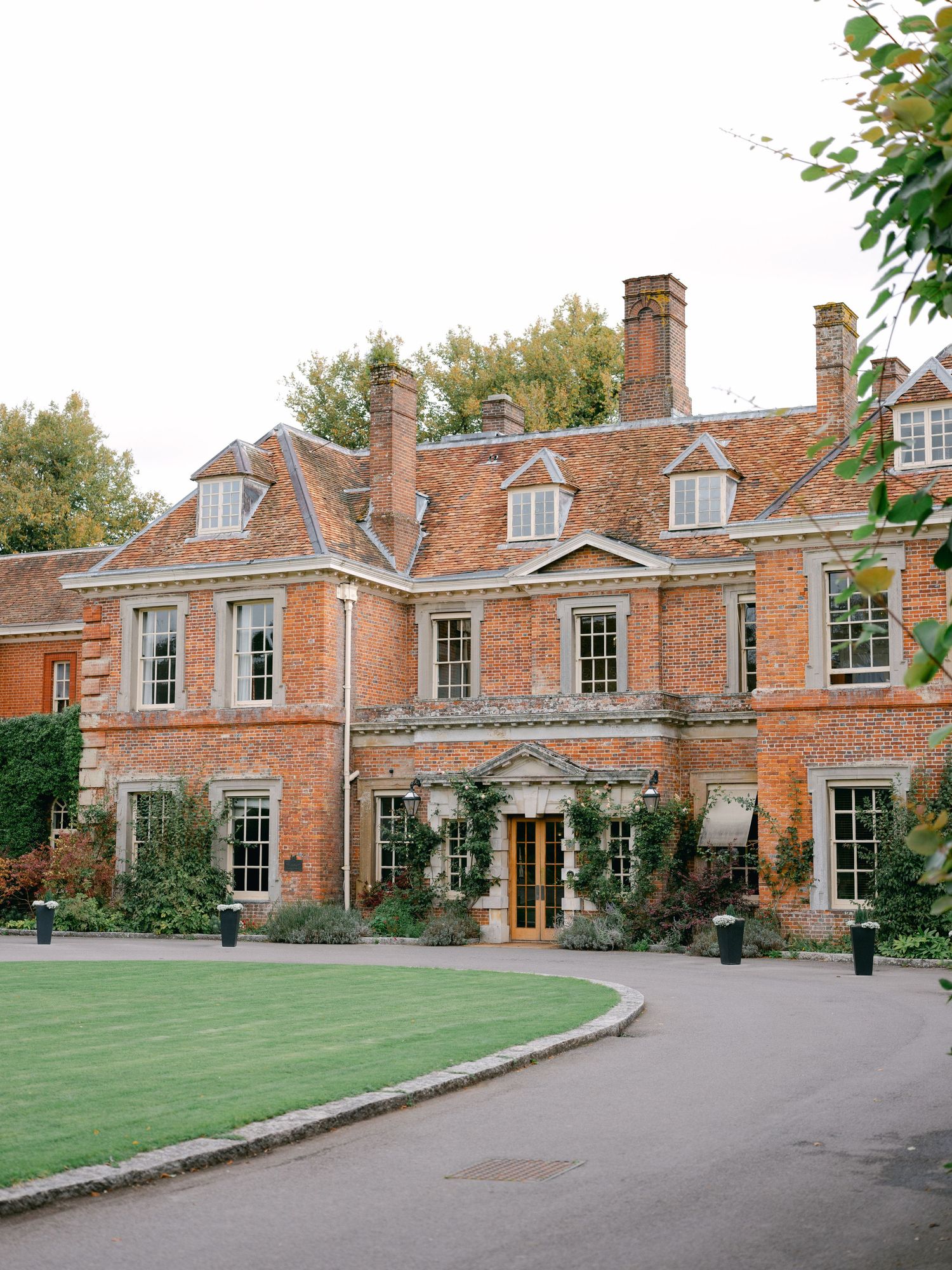 Grand red brick English manor house with dormer windows, ivy-covered walls, and manicured lawn under overcast skies.