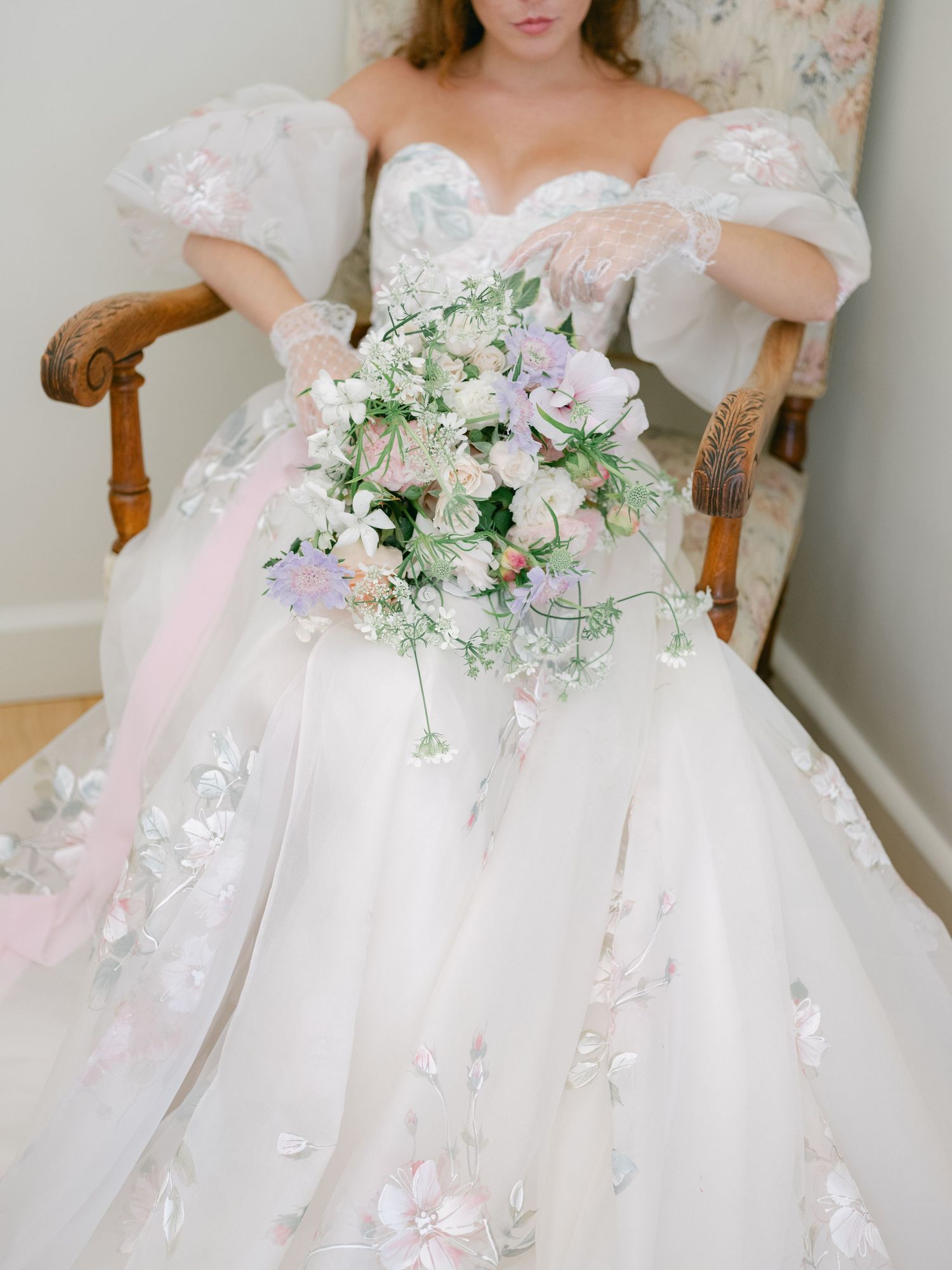 Bride in floral ball gown with puff sleeves holds cascading pastel bouquet while seated in vintage floral chair.