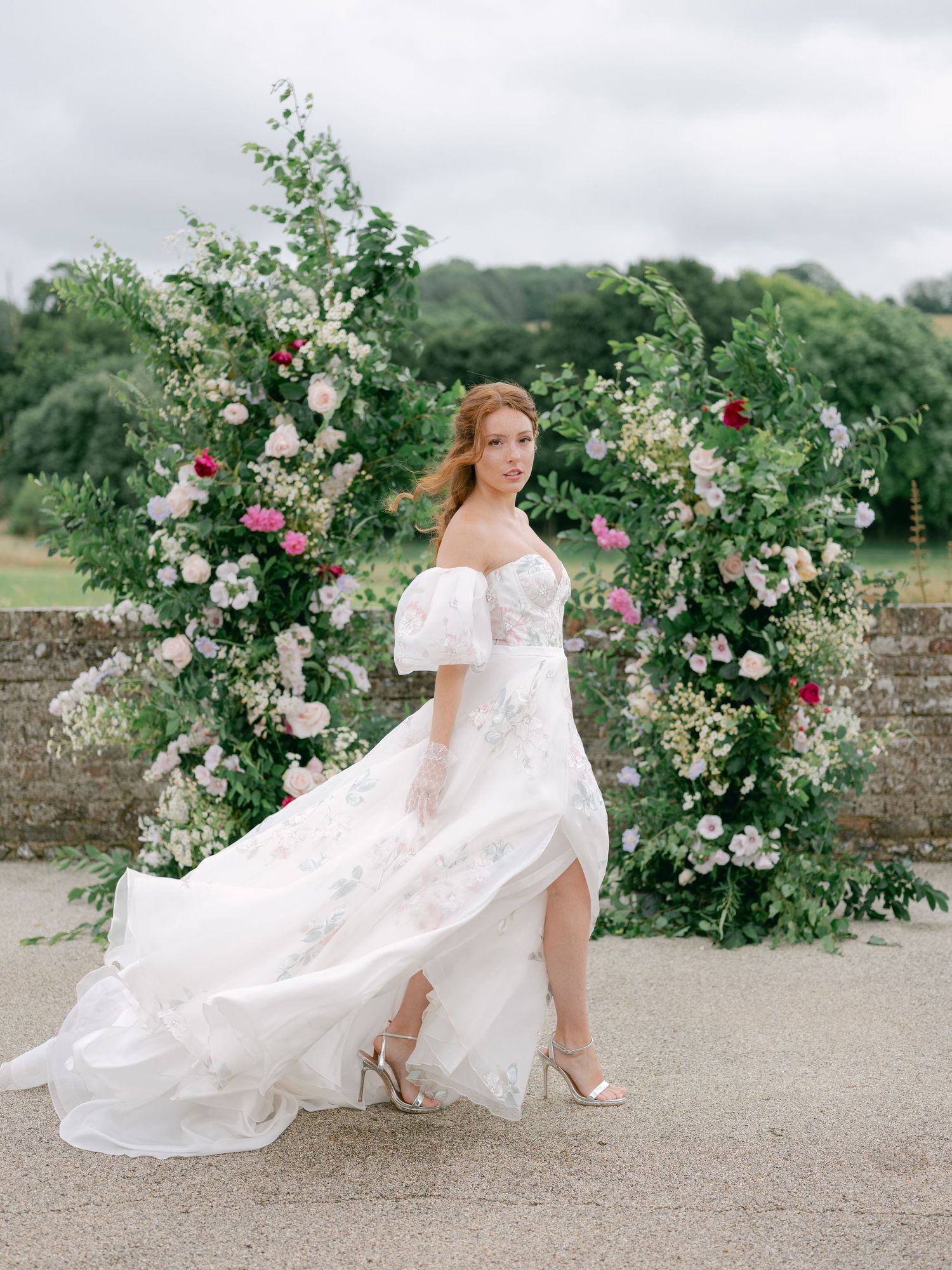 Bride in off-shoulder floral wedding gown poses before lush green floral arch with pink roses outdoors.