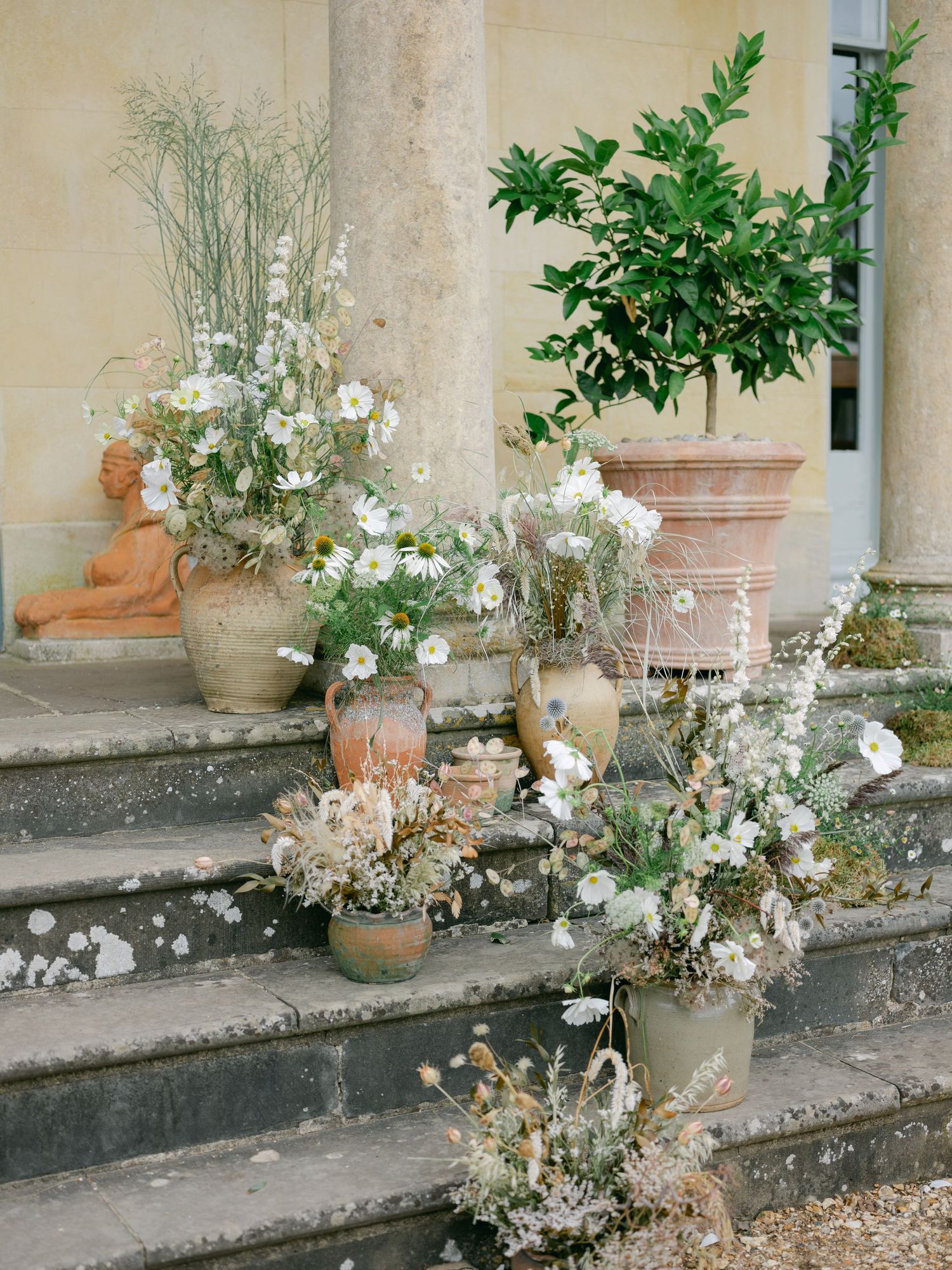 Rustic terracotta pots with white wildflowers and greenery arranged on stone steps of an elegant estate.