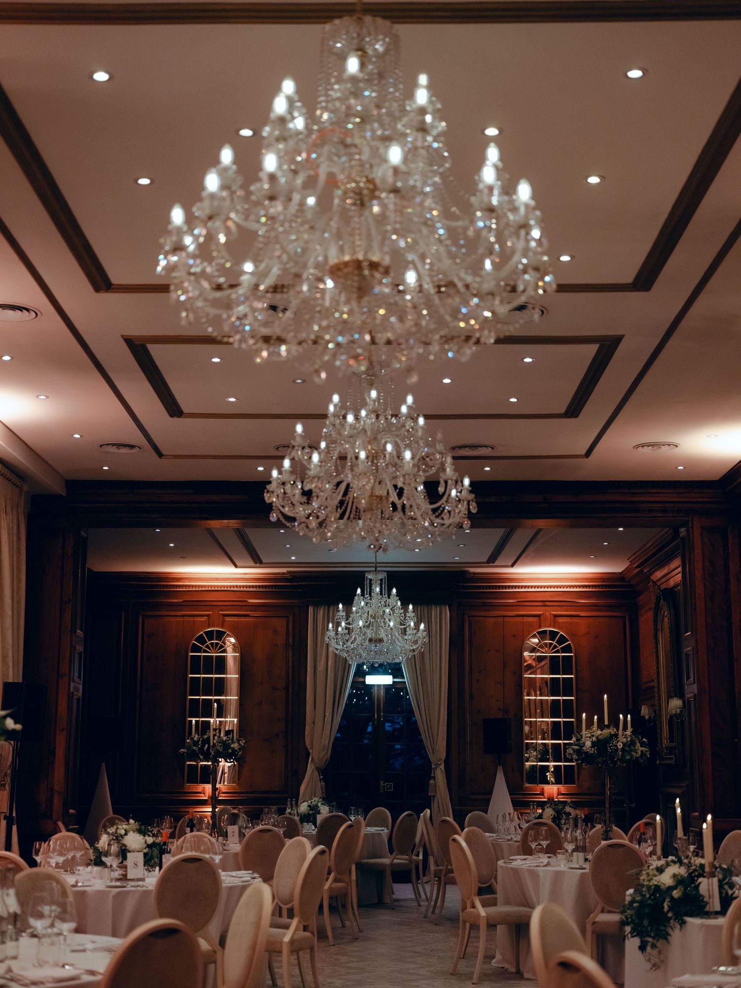 Elegant ballroom with crystal chandeliers and dark curtains set up for an evening reception with round tables and chairs.