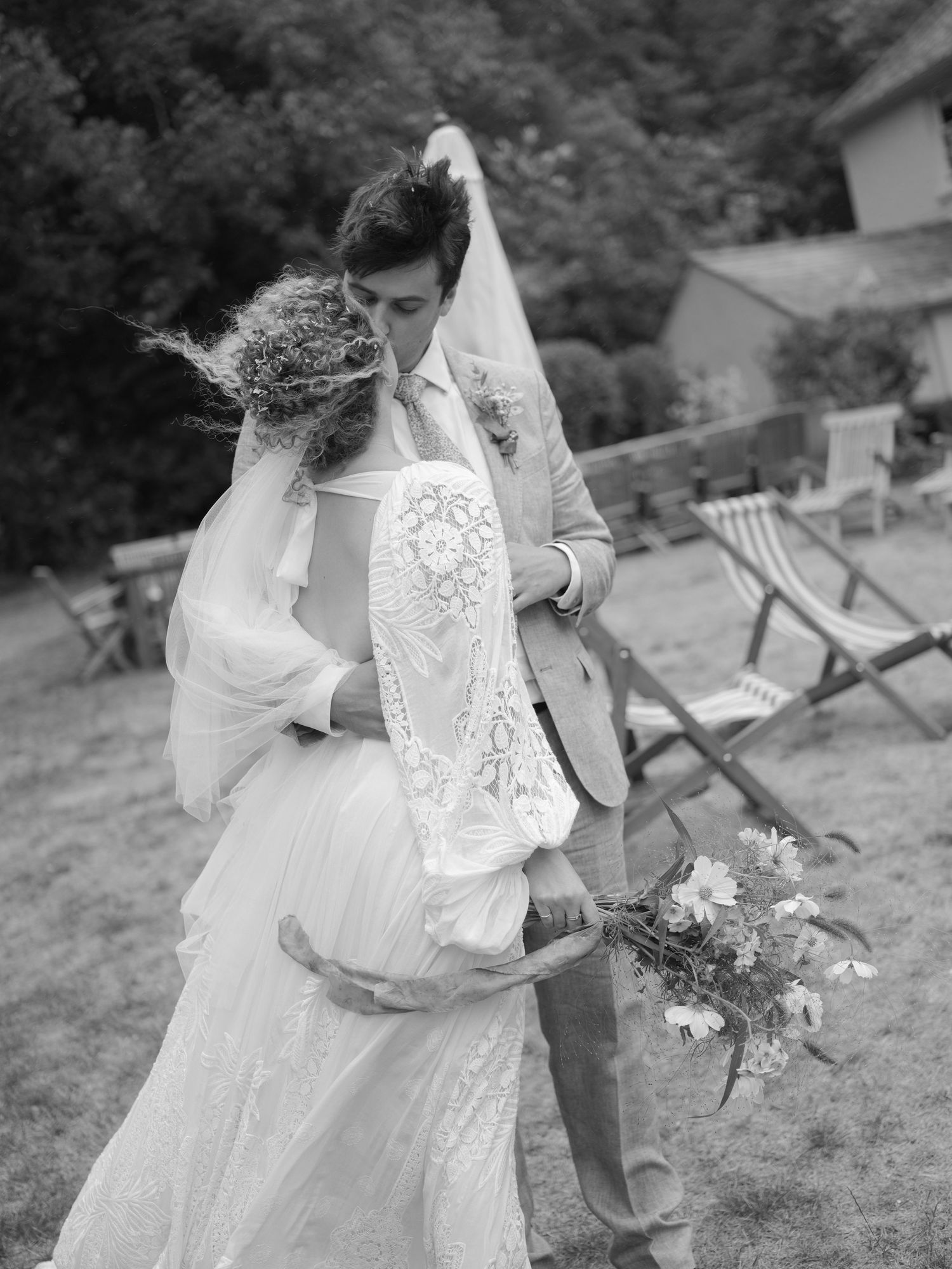 Black and white photo of a bride and groom embracing outdoors, bride holding wildflowers, veil blowing in the wind.