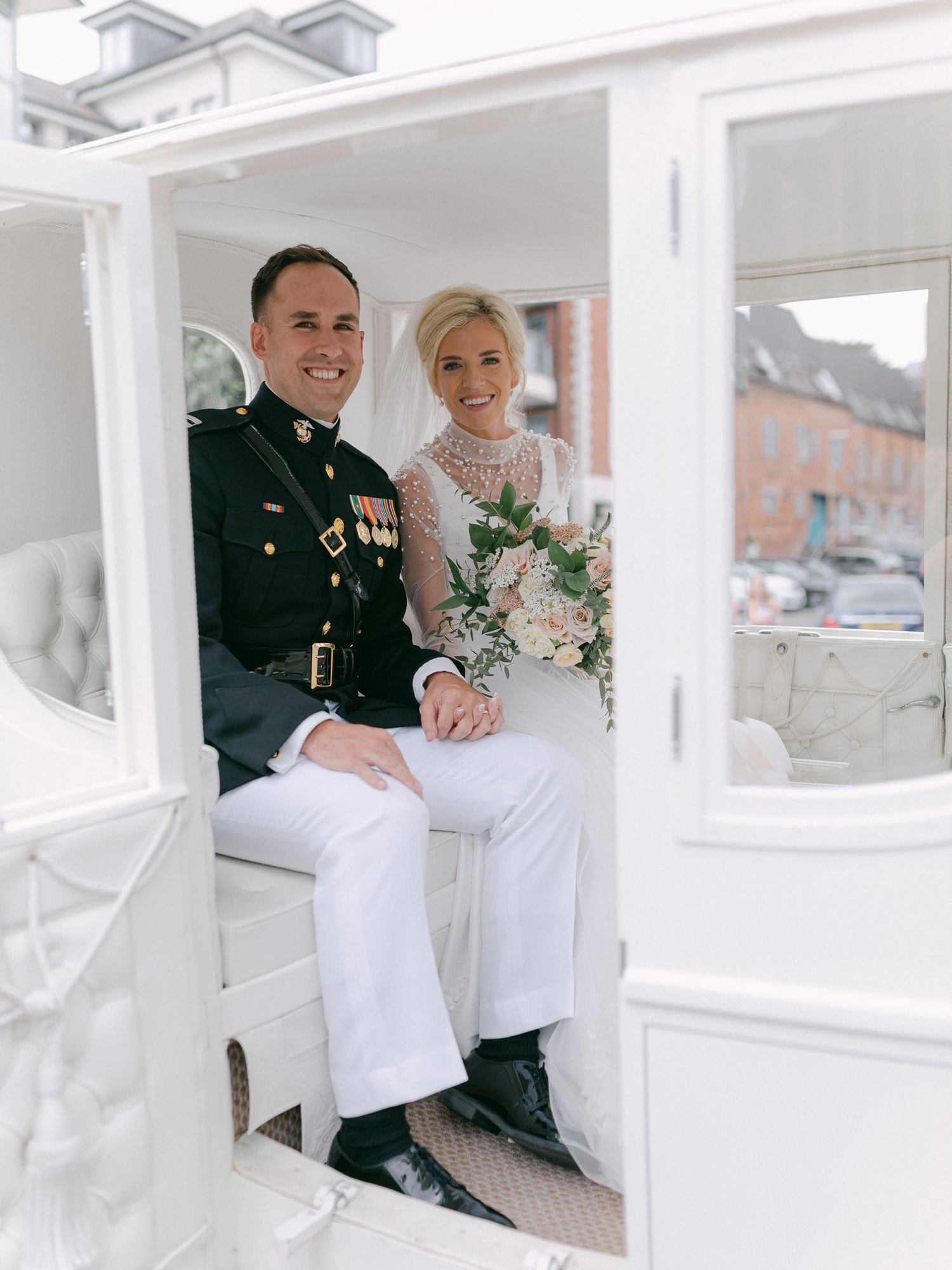 Military groom in dress uniform and bride in white lace gown holding bouquet sitting in elegant white carriage on wedding day.