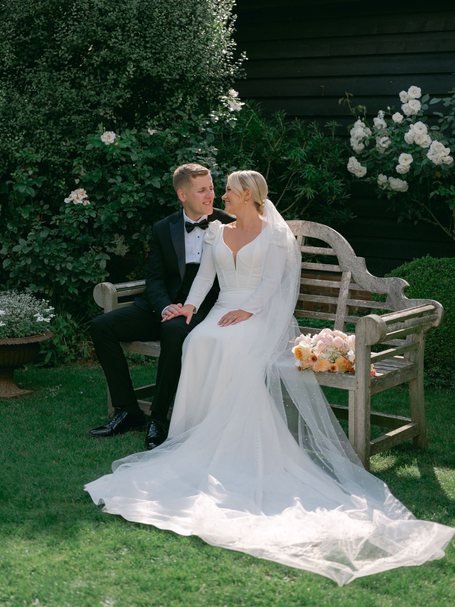 Bride and groom sit on a wooden garden bench surrounded by white roses, sharing a romantic gaze on their wedding day.