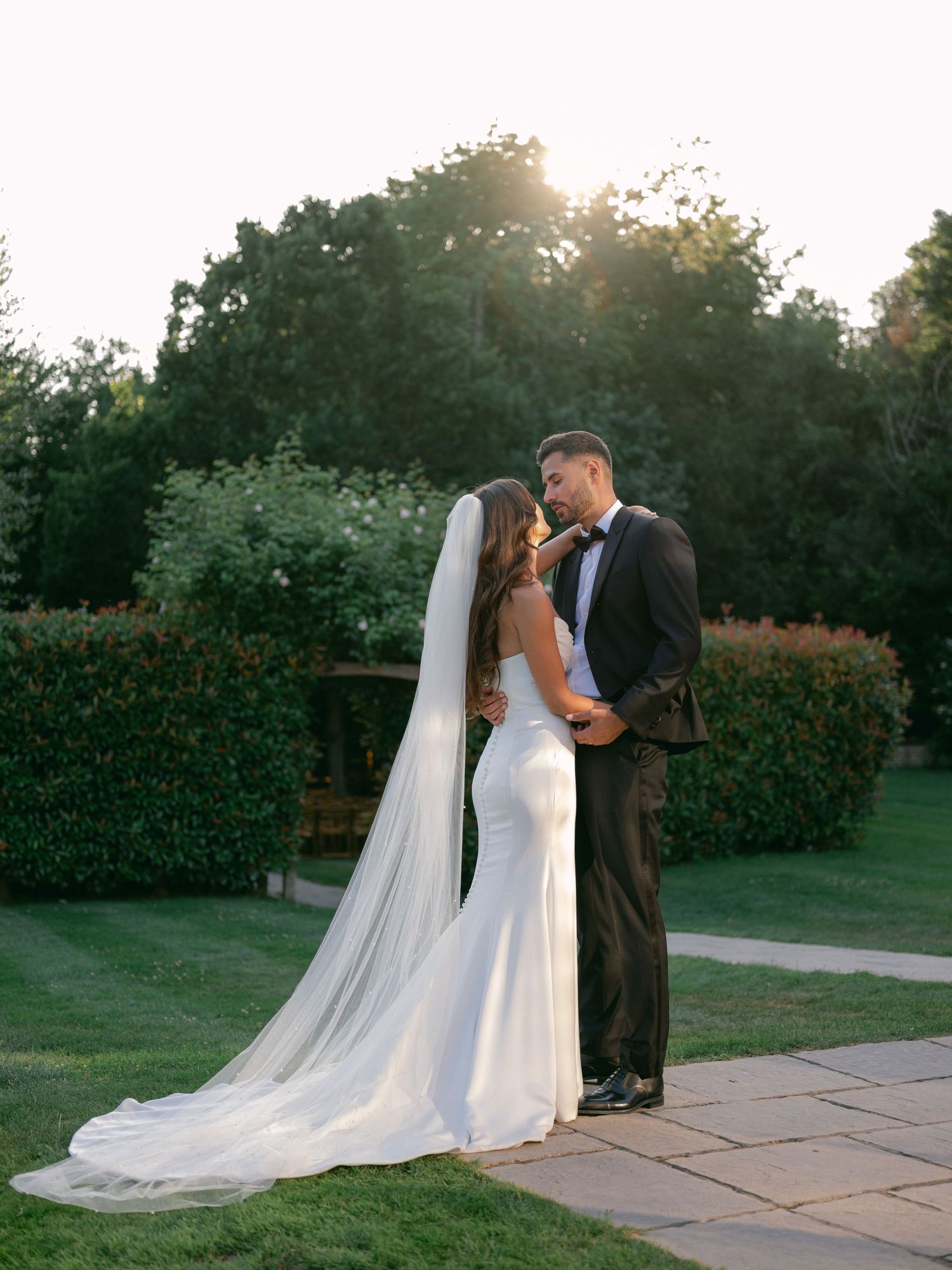 A couple shares a romantic kiss at sunset on a stone patio with a long flowing veil and lush garden backdrop.