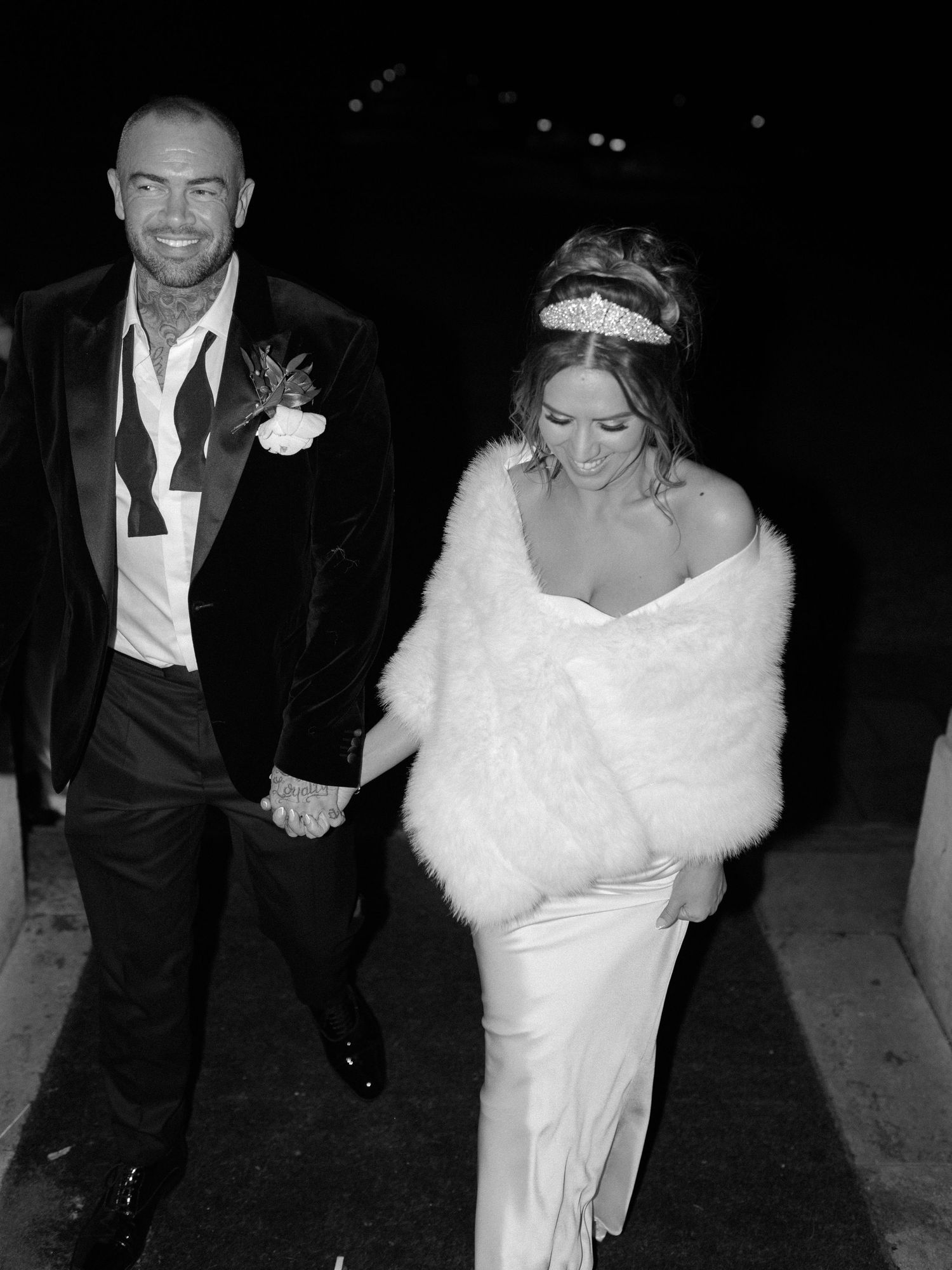 A black and white photo of a couple at a formal event, with the bride wearing a white gown and fur wrap.