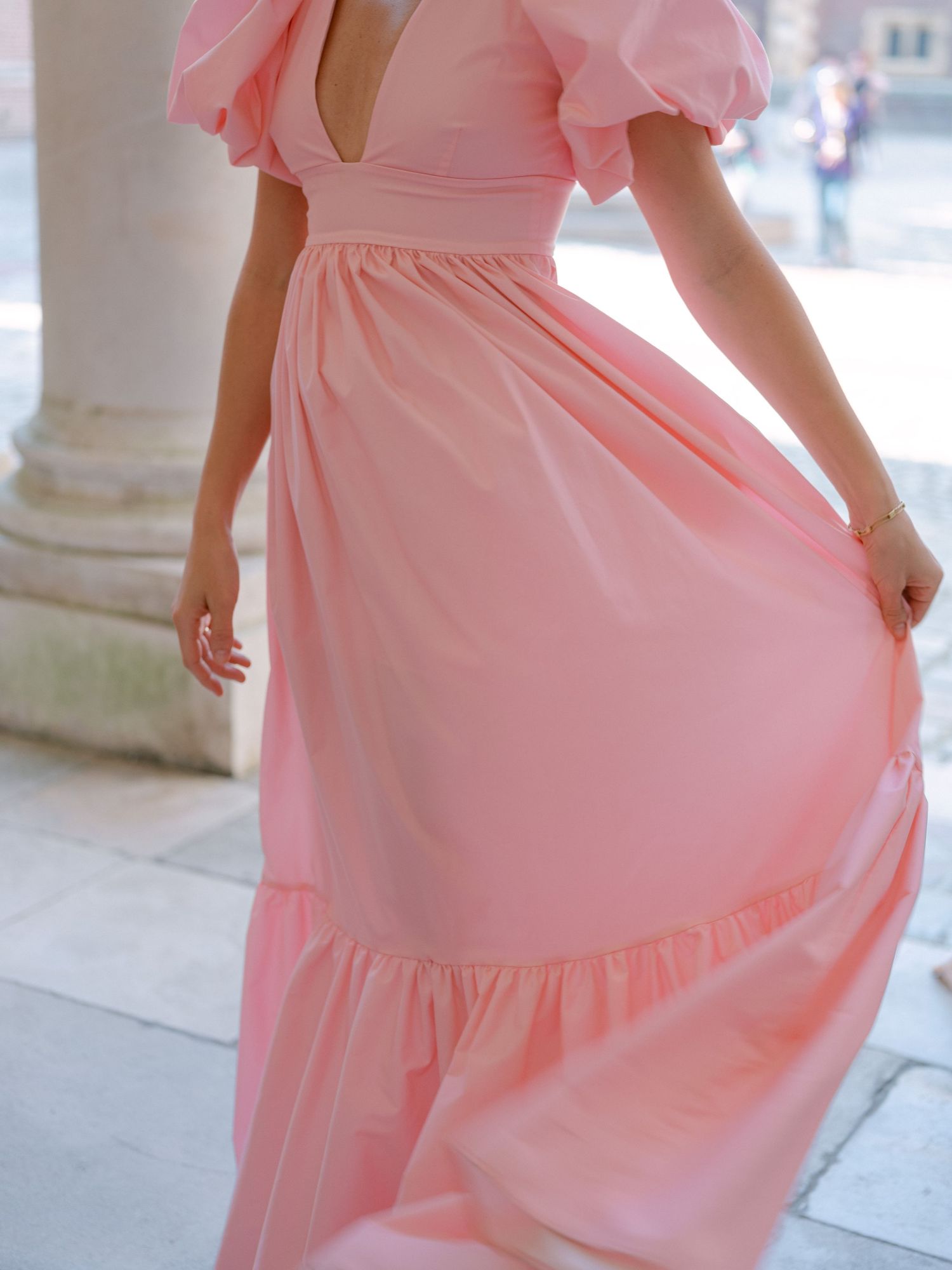 A woman wearing a pink maxi dress with puff sleeves and a deep V-neckline, standing near a stone column.
