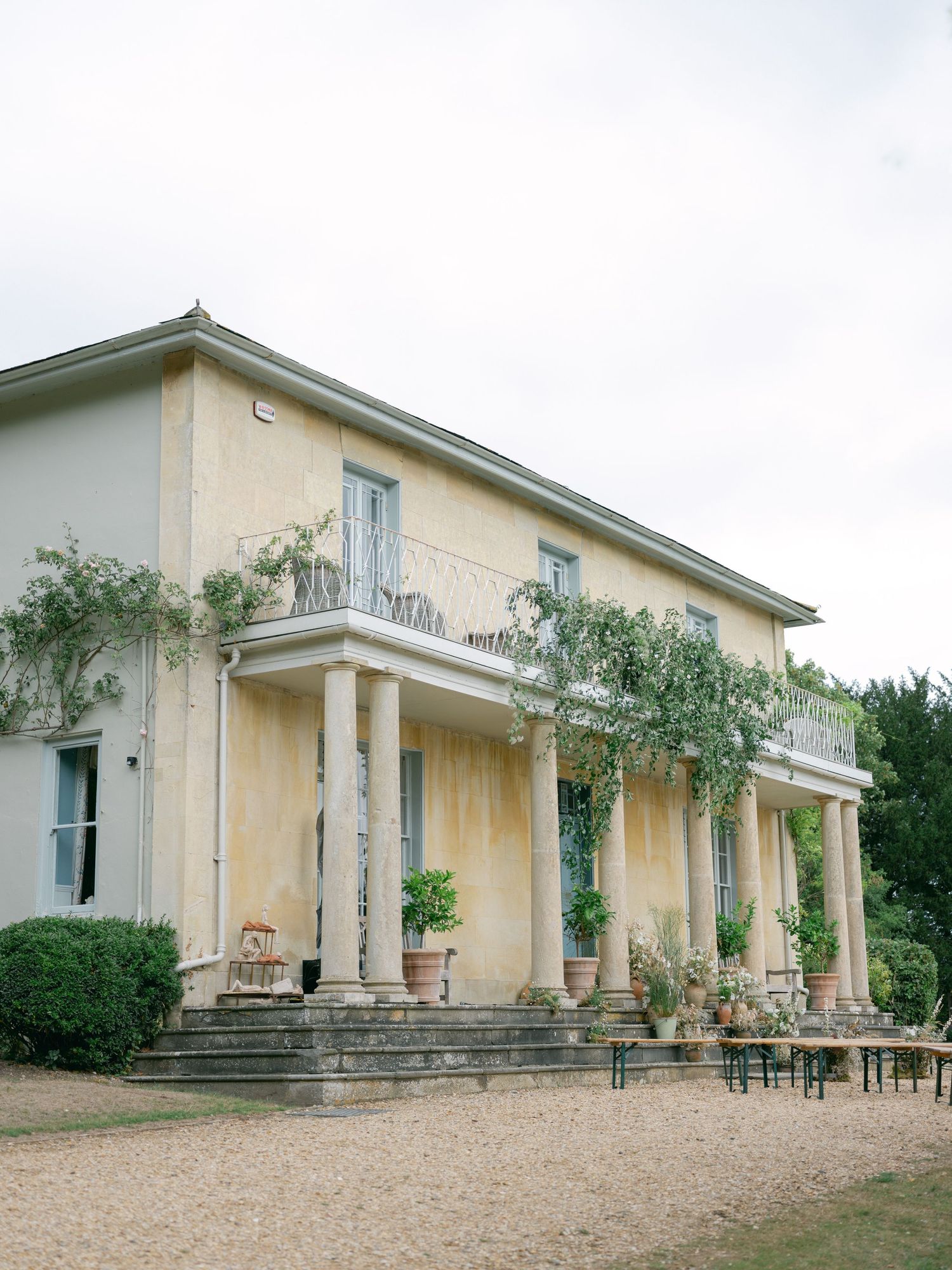Elegant neoclassical villa with cream columns and balcony features gravel driveway and potted plants on a tranquil estate.