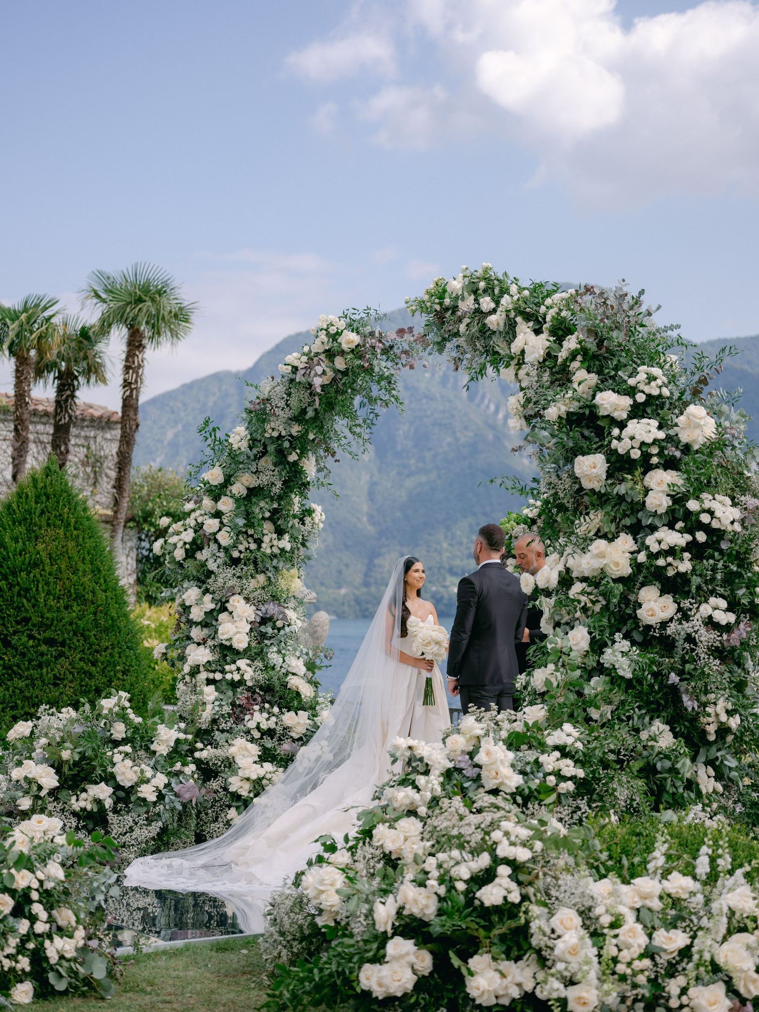 Bride and groom exchange vows under a lush floral arch with white blooms, set against mountain and lake views.