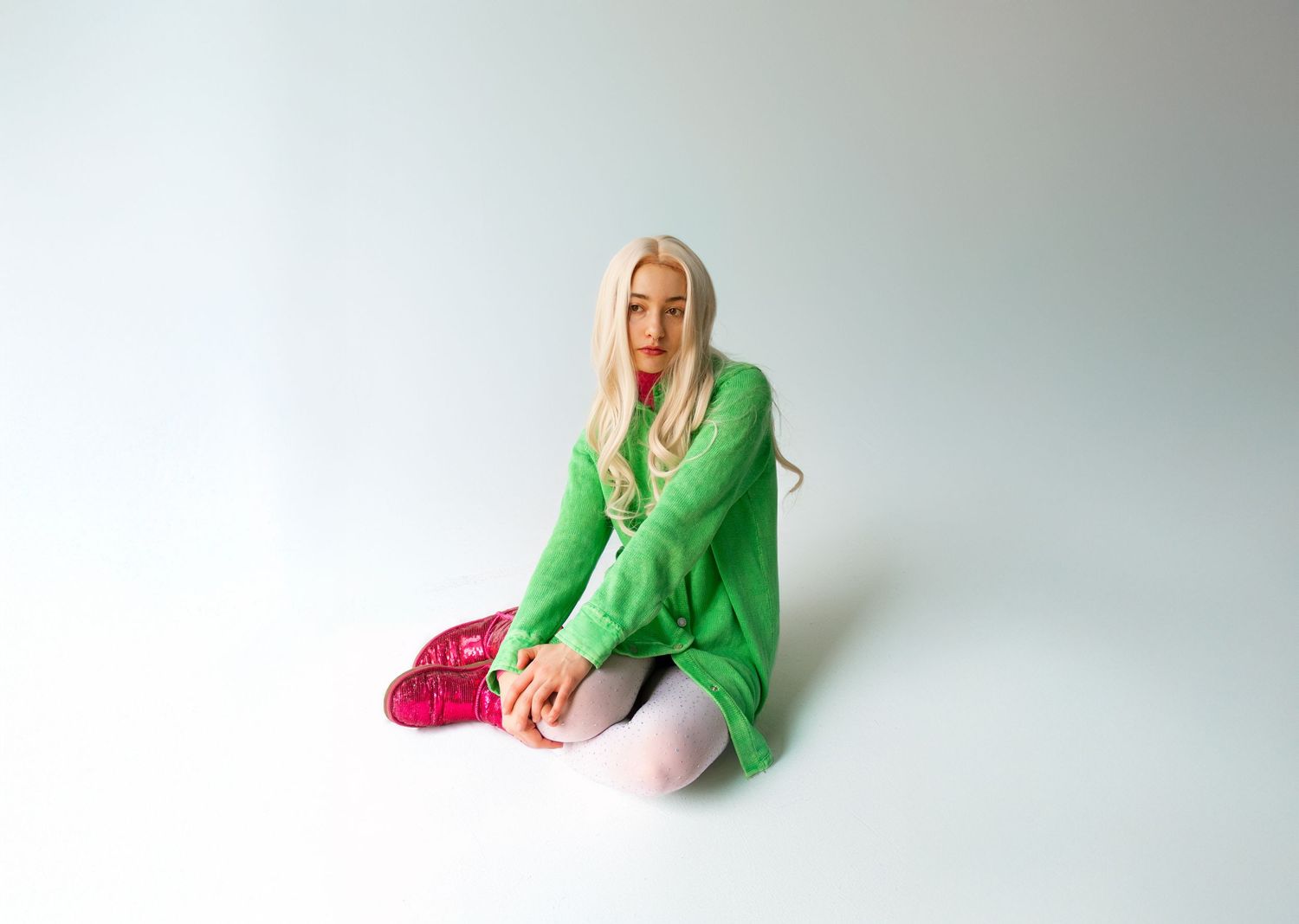 Blonde woman in green oversized sweater, pink shoes, and white tights sitting on a white studio floor.