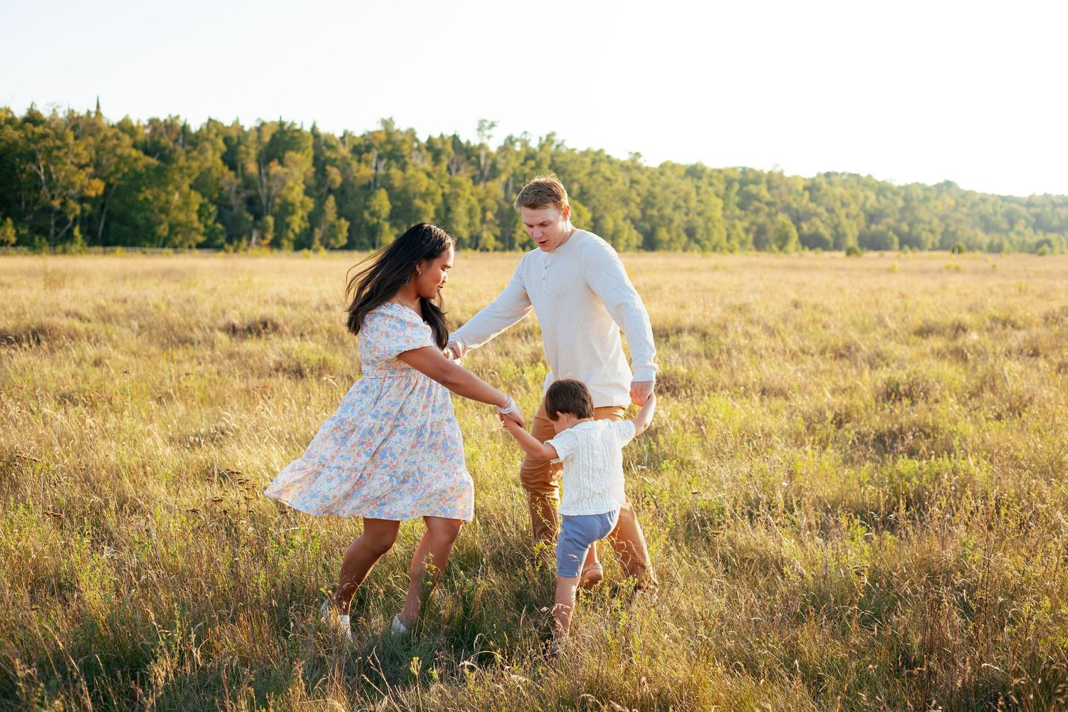 Family of three playing in a sunlit golden meadow with lush green trees in the background on a warm sunny day.