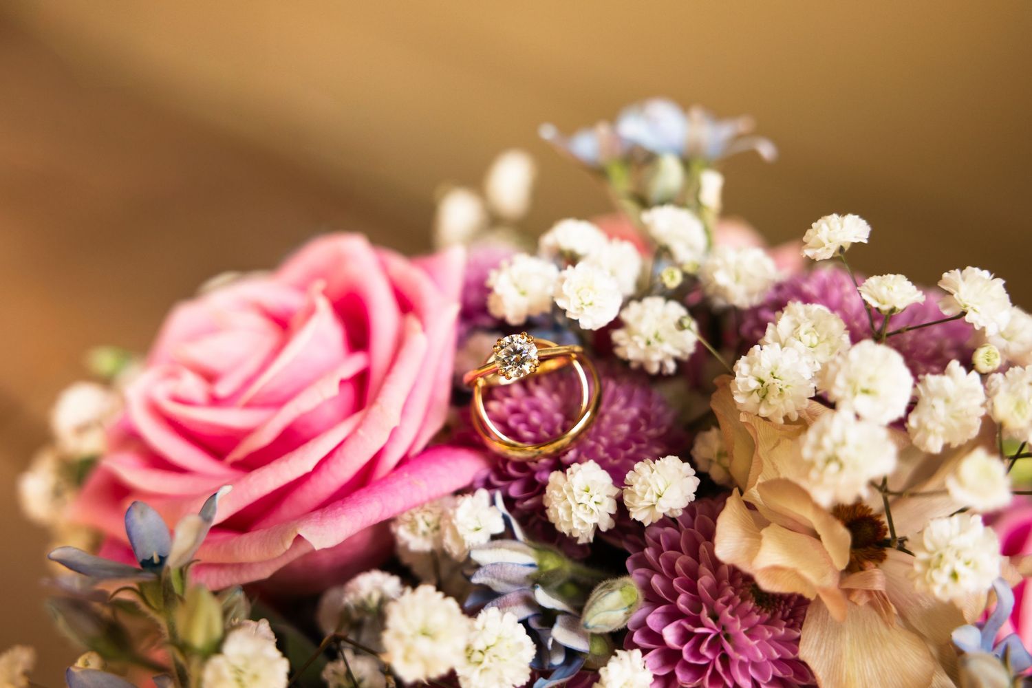 Elegant floral arrangement featuring pink roses, white baby's breath, and purple blooms against a warm brown background.