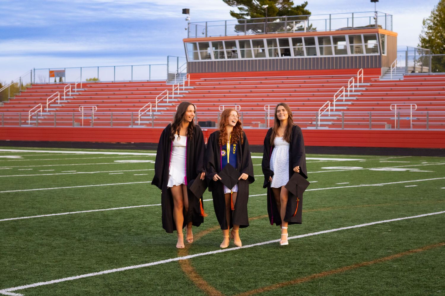 Three young women in graduation gowns walking on a football field with red bleachers in the background.