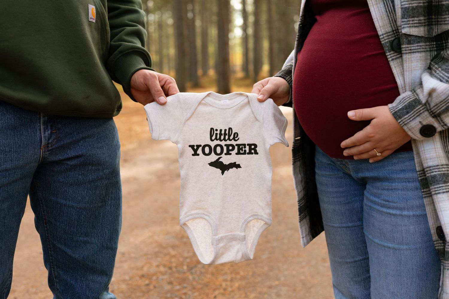 Couple holding tiny white baby onesie in autumn forest setting during maternity photoshoot.