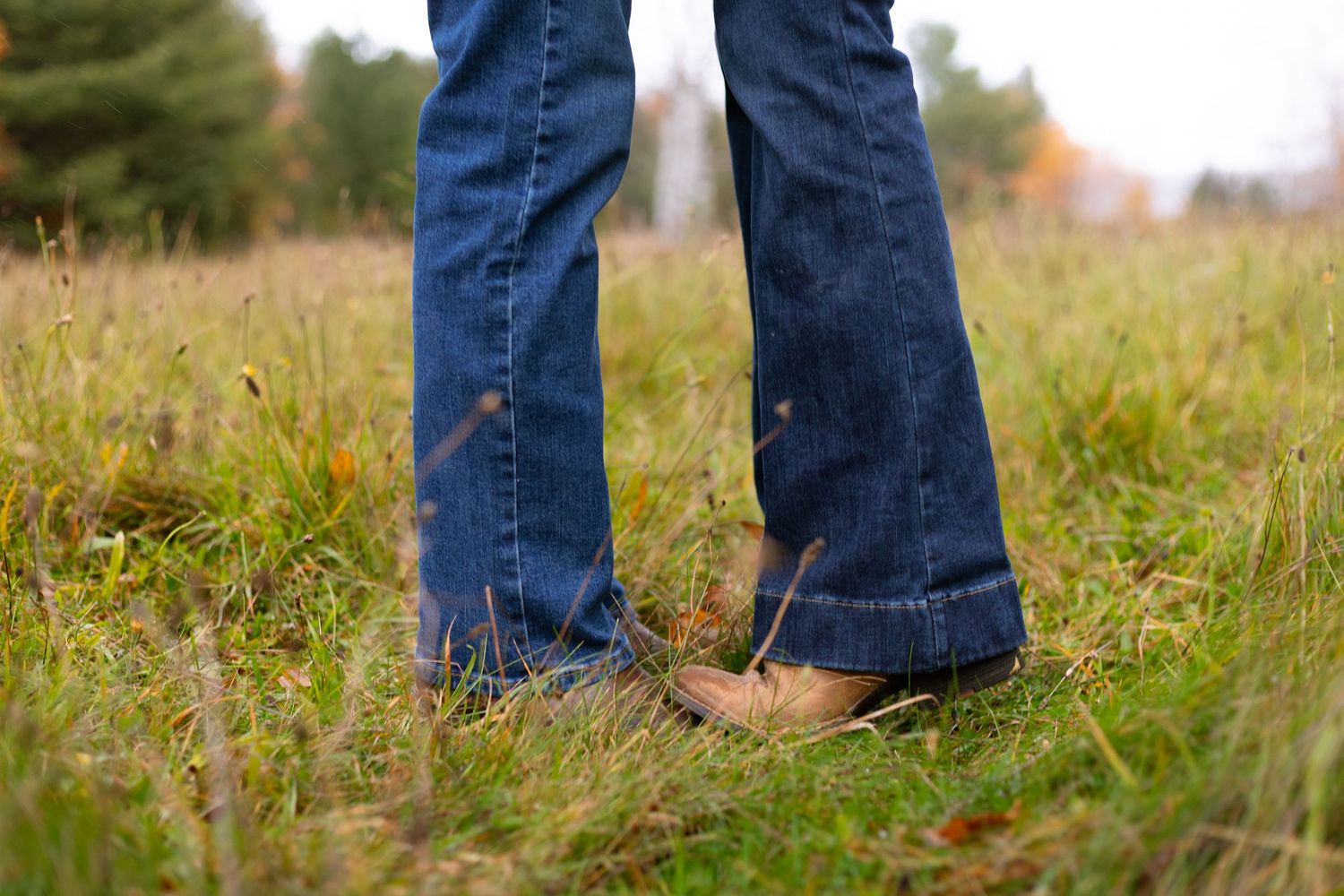Two pairs of feet in blue jeans and brown shoes stand close together in a grassy autumn field.