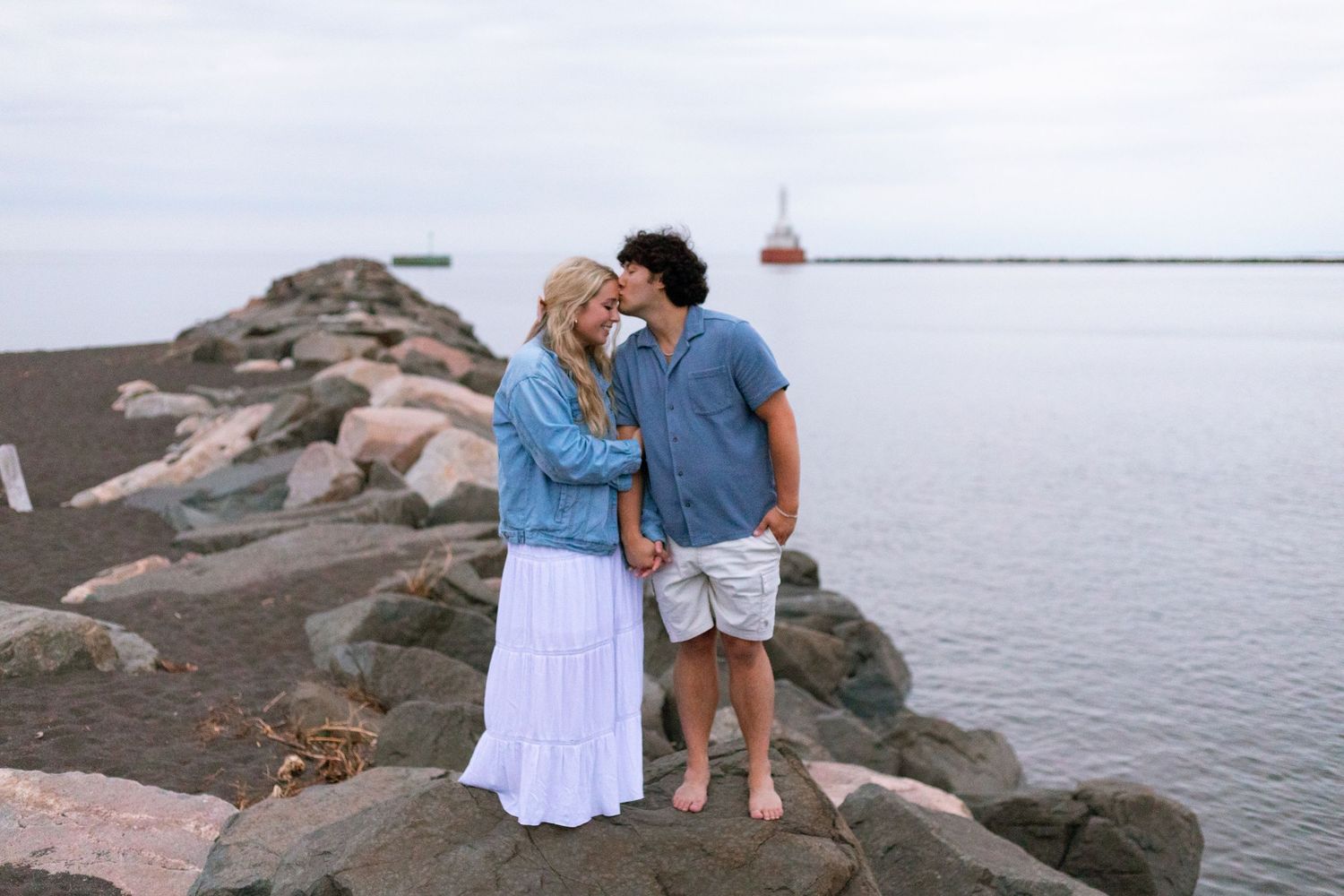 Couple standing on rocky jetty by the ocean, woman in white dress and denim jacket, man in blue shirt, lighthouse in background.