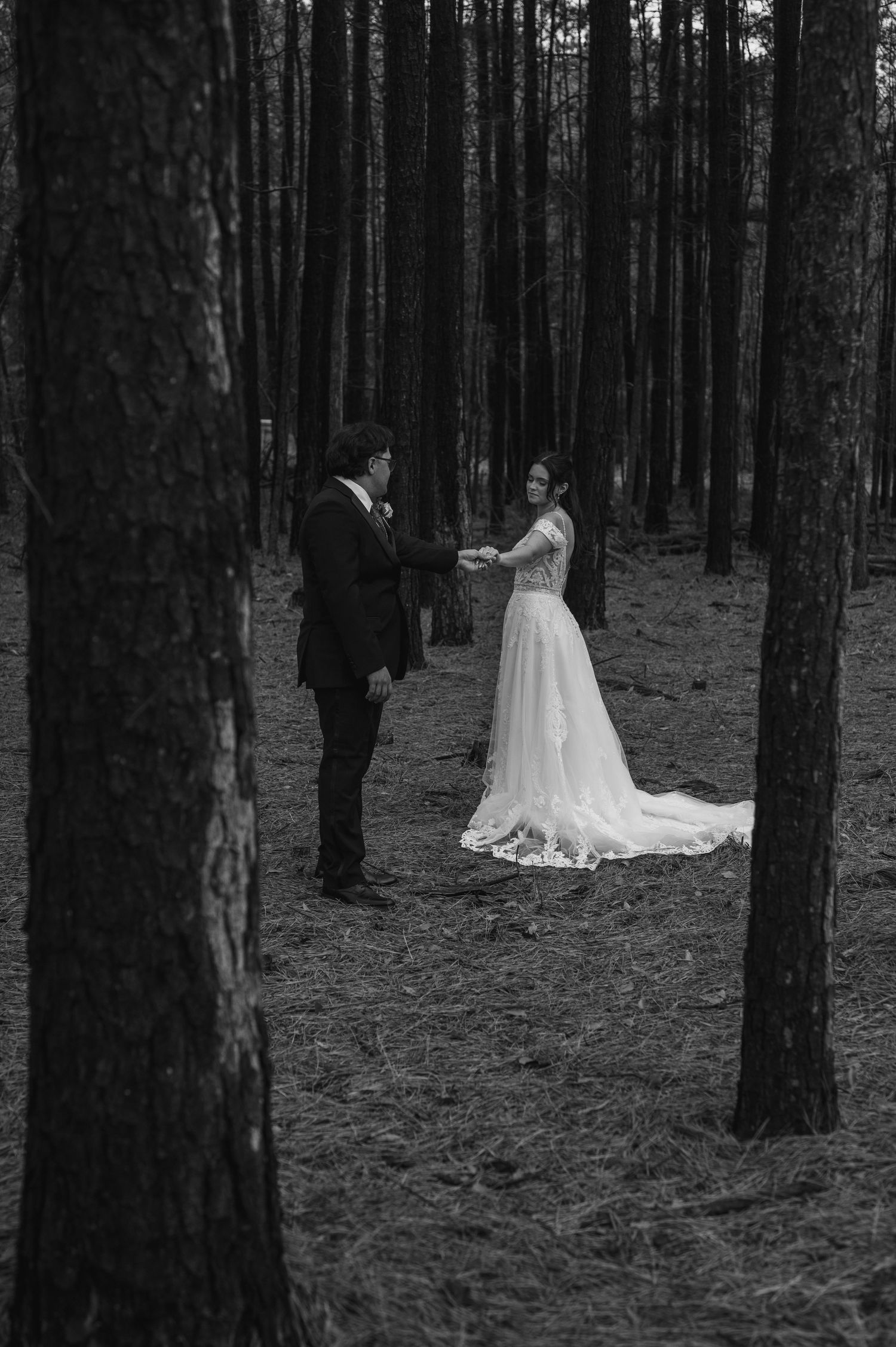 A black and white wedding photo of a couple standing together in a serene pine forest.