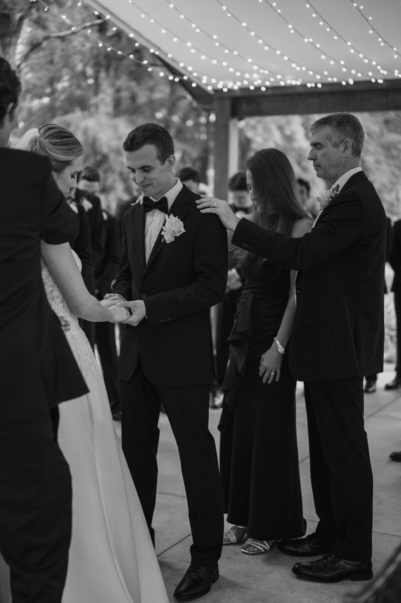 Black and white photo of a formal wedding reception under string lights with people dancing and mingling.
