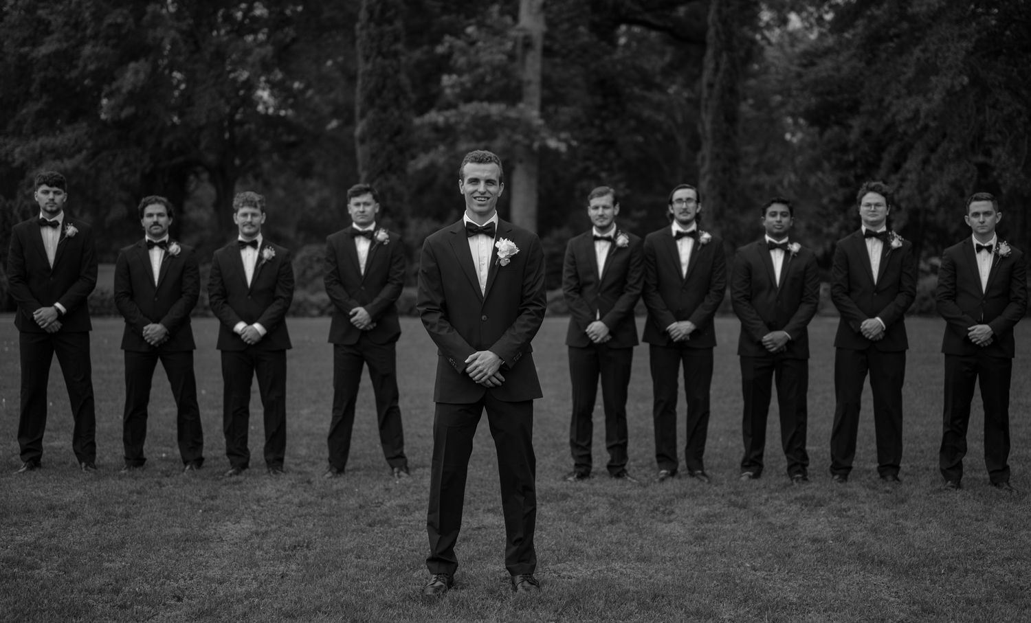 A black and white photo shows a large wedding party lined up outdoors in formal attire.