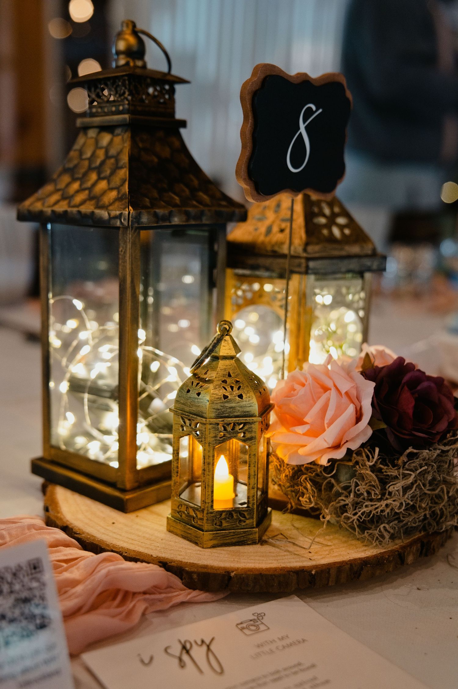 Rustic wedding centerpiece with brass lanterns, fairy lights, pink roses, and a chalkboard table number on a wood slice.