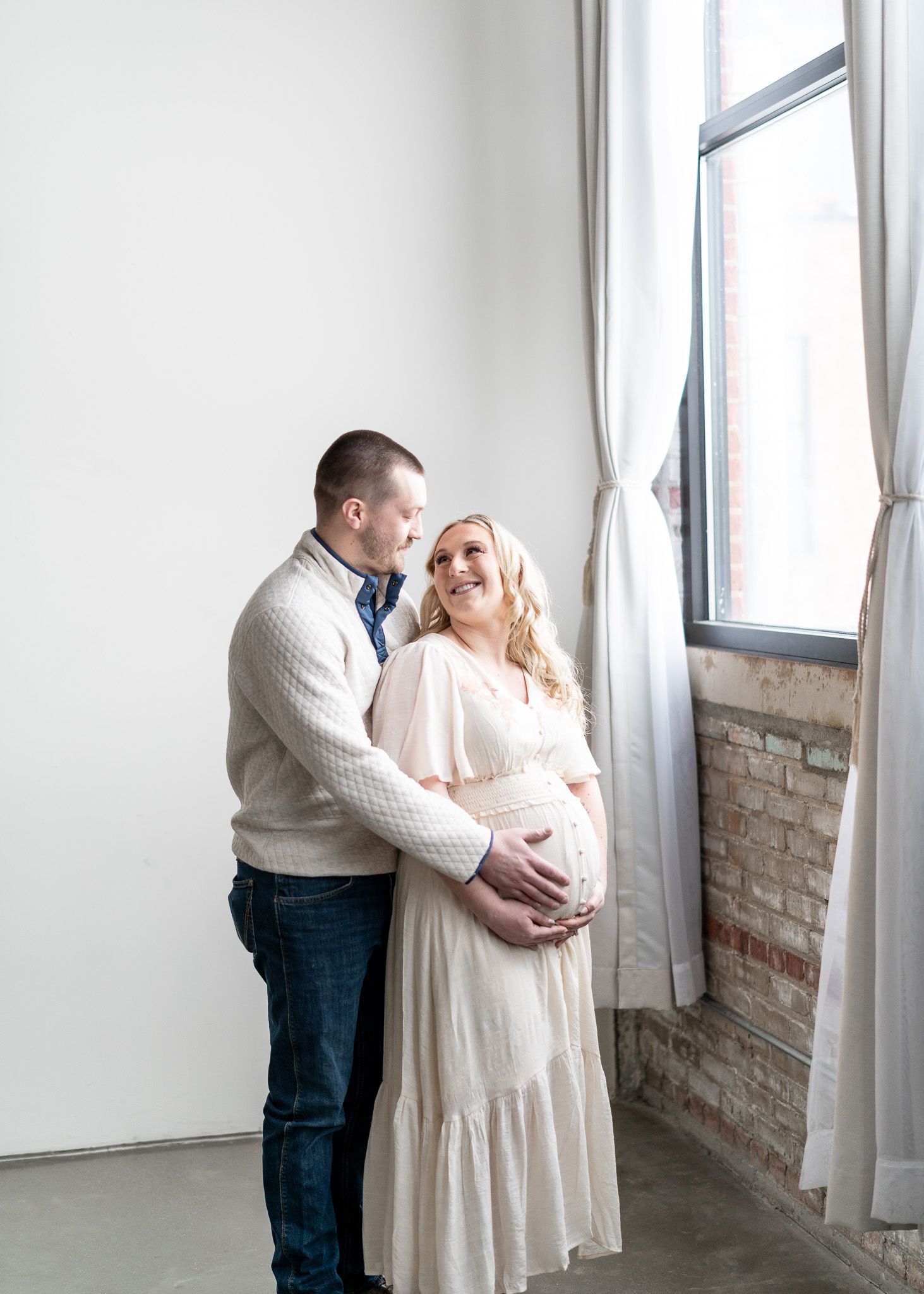 A couple in casual attire shares tender moments in a bright industrial-style room with exposed brick walls and white curtains.