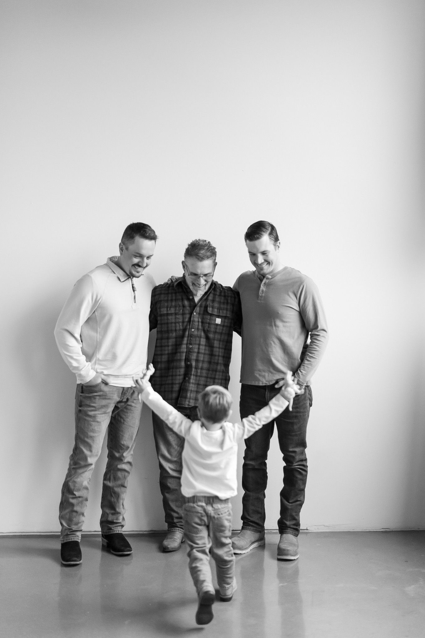 Black and white portrait of three people in casual attire holding hands with a small child in a playful pose.