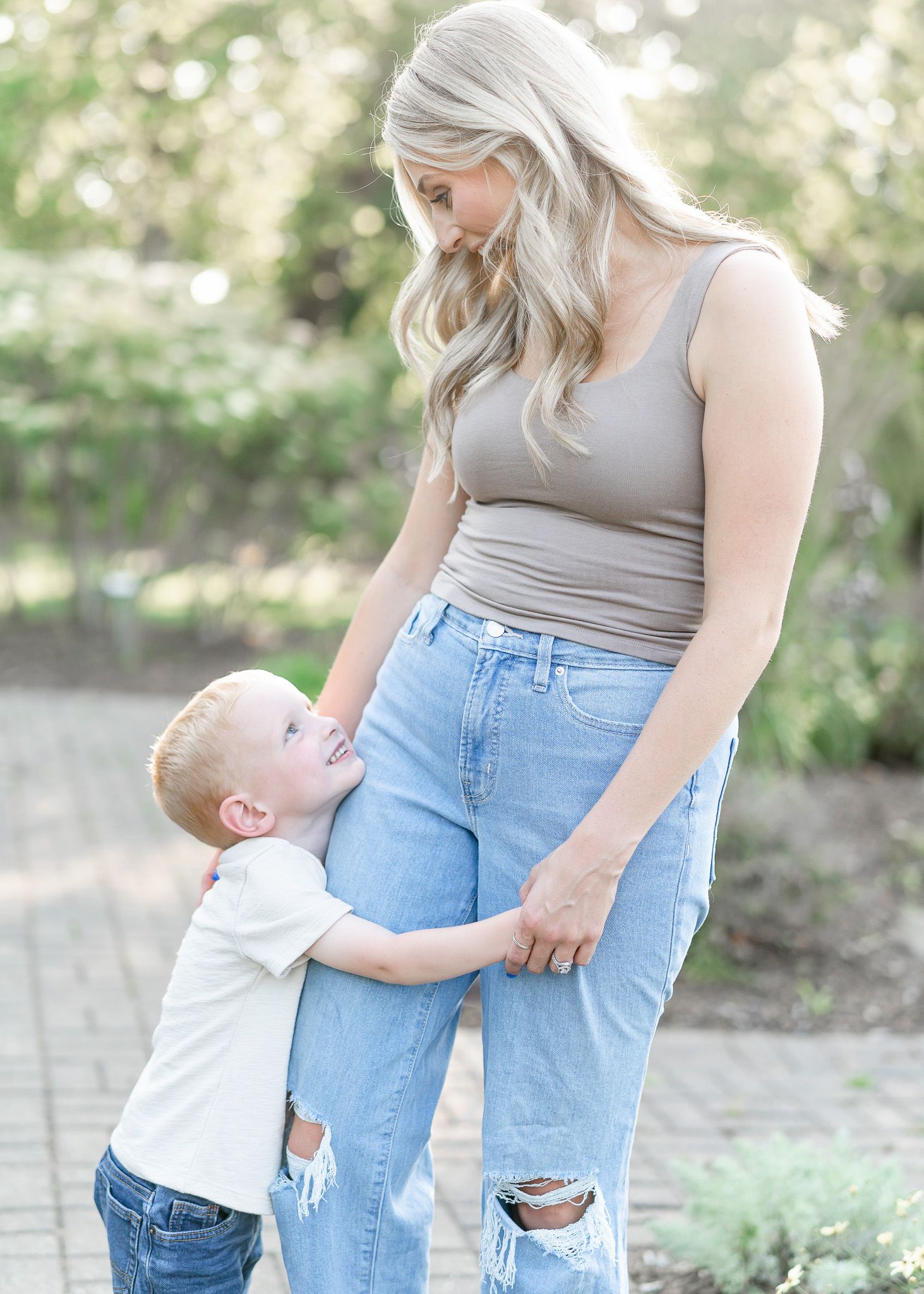 A young child in white hugs the leg of someone wearing a gray tank top and distressed light blue jeans outdoors.