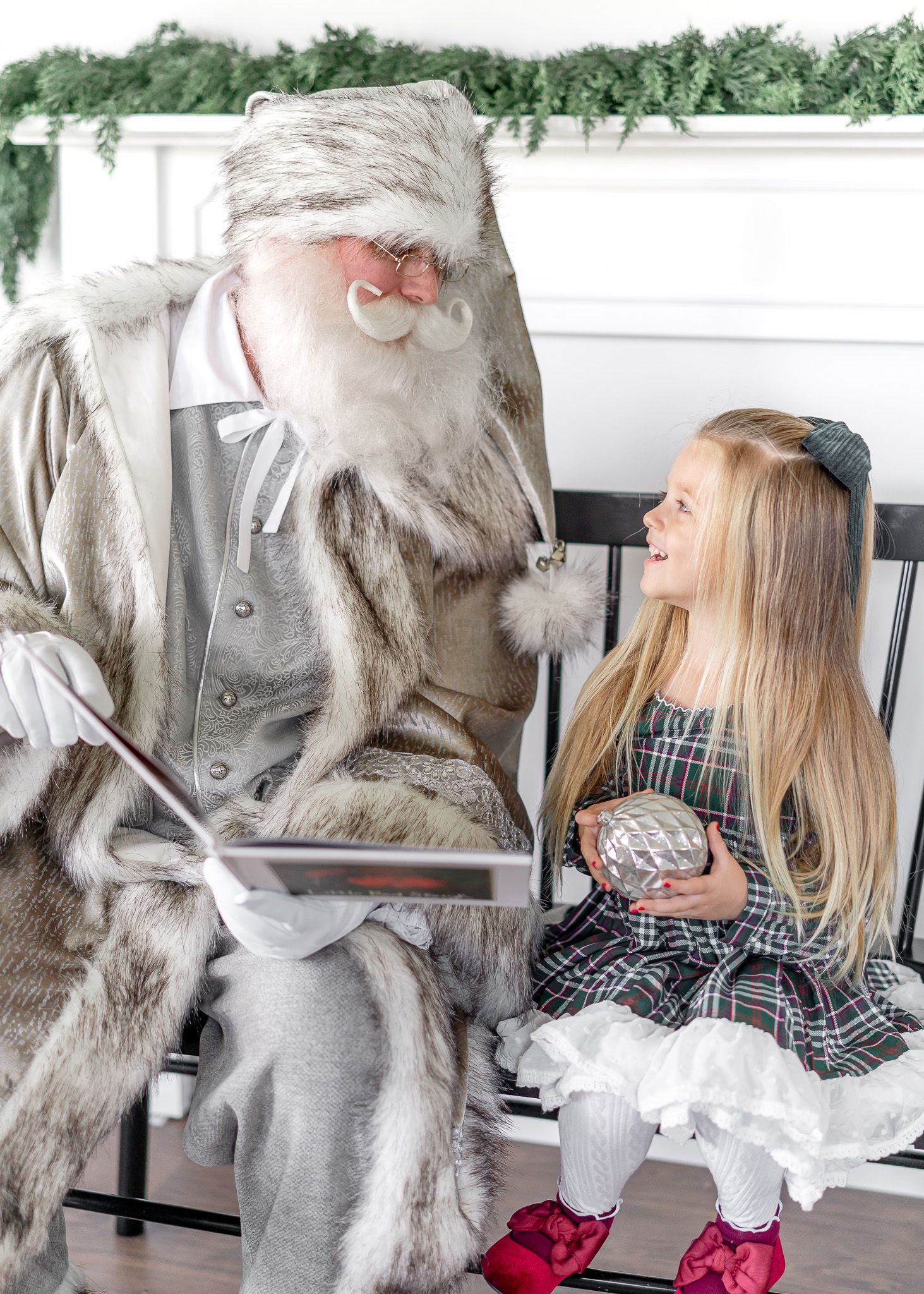 A magical holiday scene with Santa Claus in elegant grey attire sitting with a young child in a festive dress.