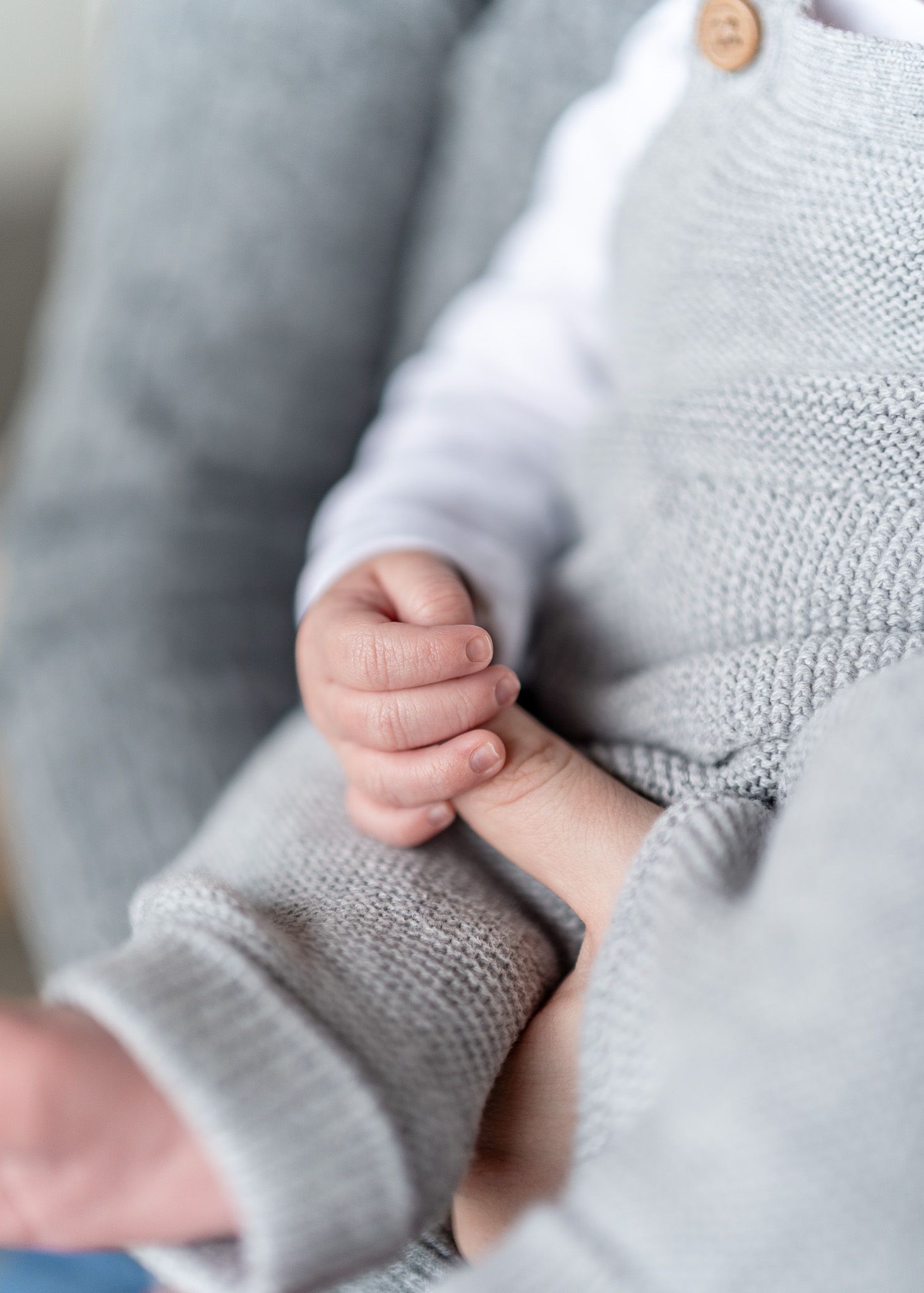 A tiny baby hand gently grasps an adult's finger while wearing a soft gray knit sweater.