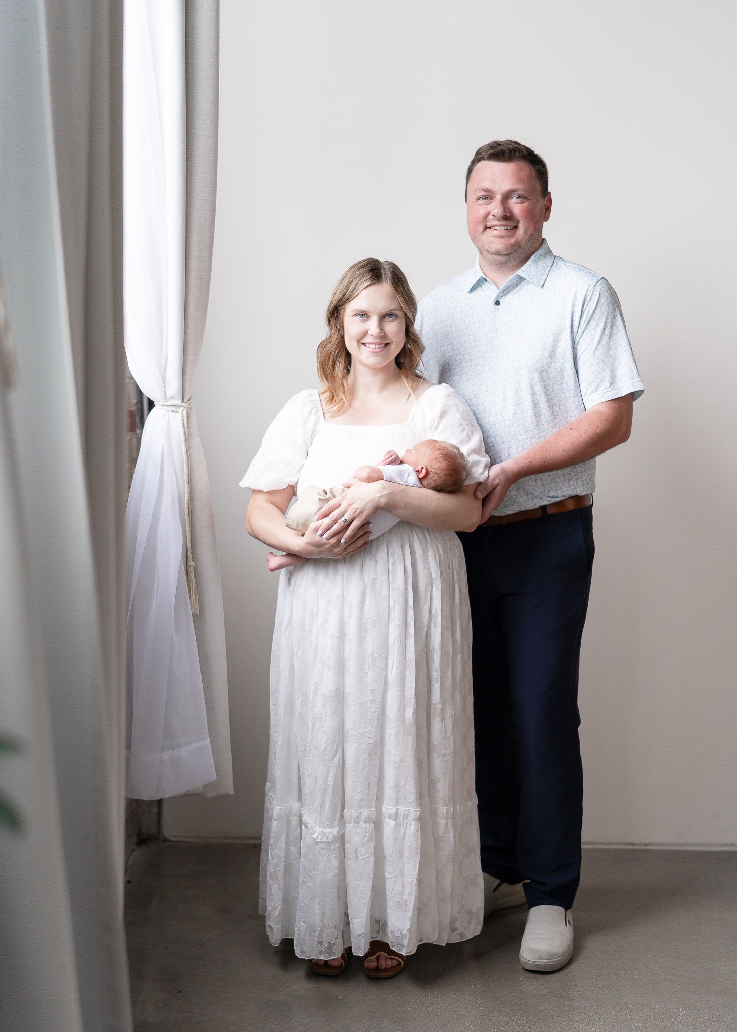 A couple in white clothing poses together in a studio setting with white curtains in the background.
