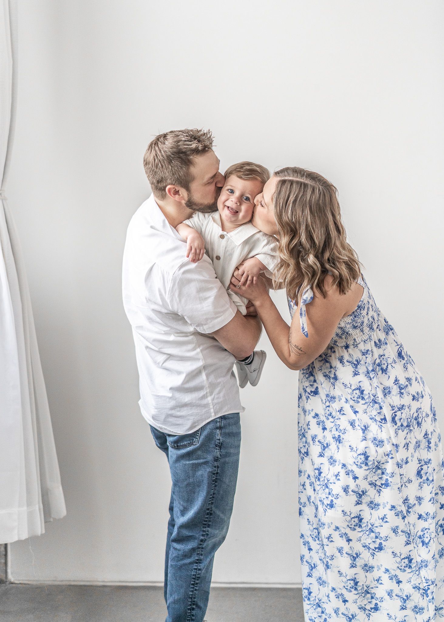 Parents in white and blue floral dress share tender moments with baby in white against minimalist studio backdrop.