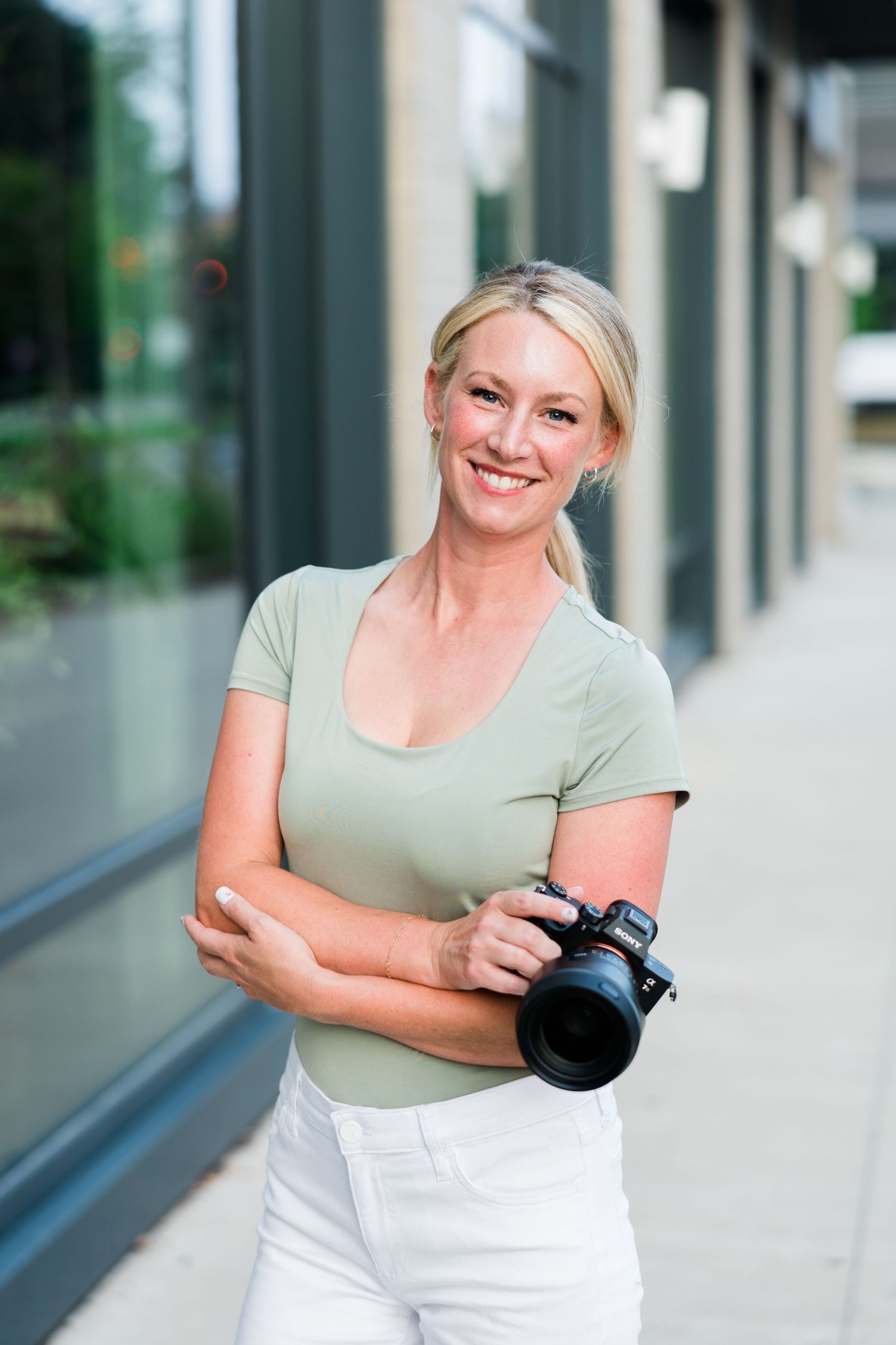 Smiling blonde woman holding a DSLR camera outdoors, wearing a green top and white jeans near a modern building.