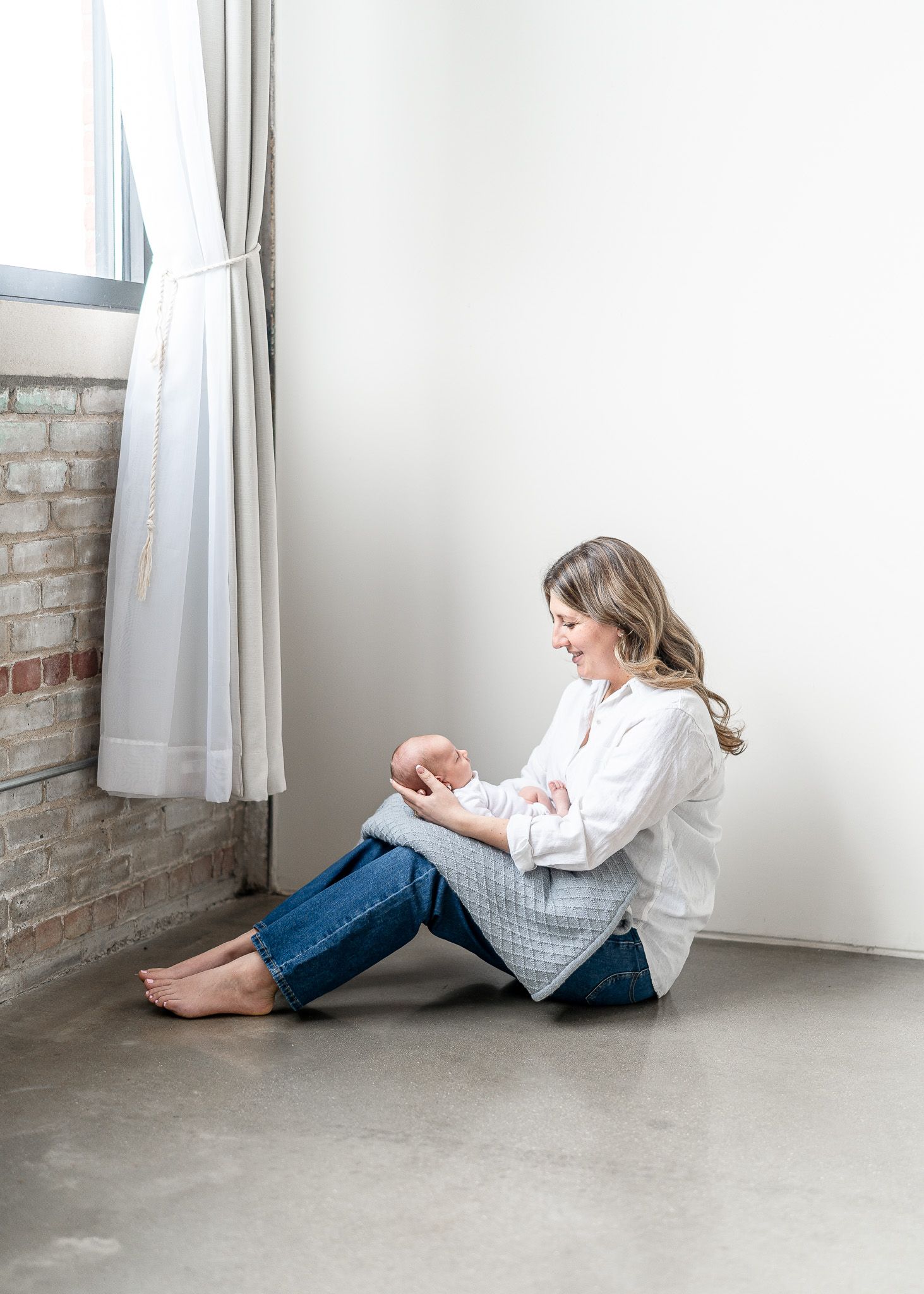 A person in a white sweater and jeans sits curled up on a white floor against a white wall with exposed brick.