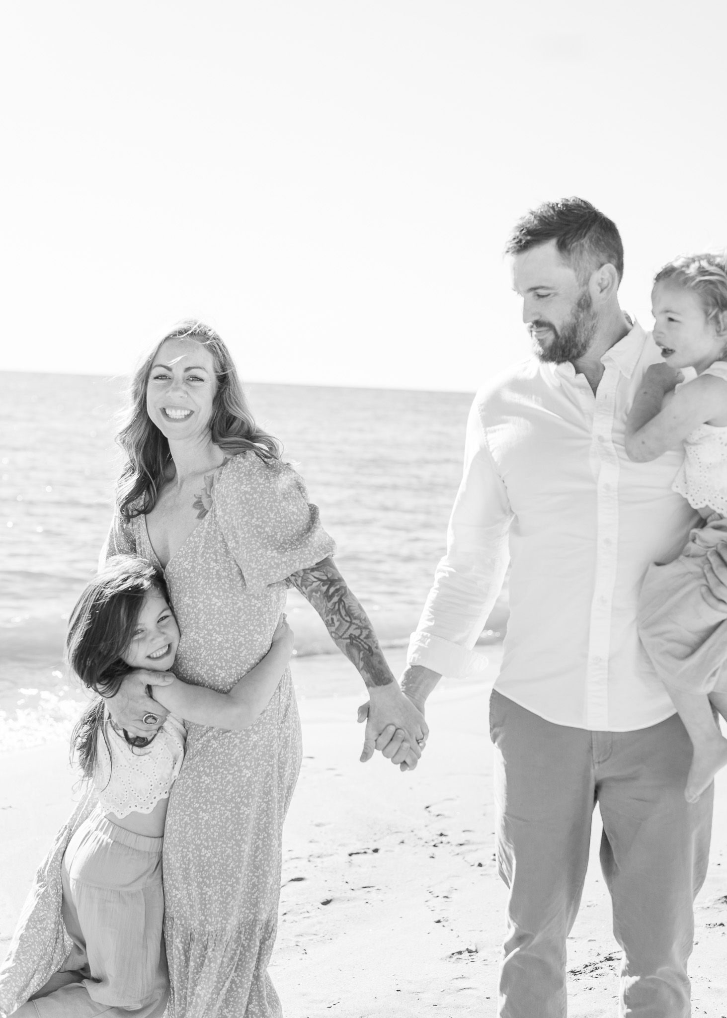 A joyful family shares intimate moments together while walking along a sunny beach in casual white outfits.