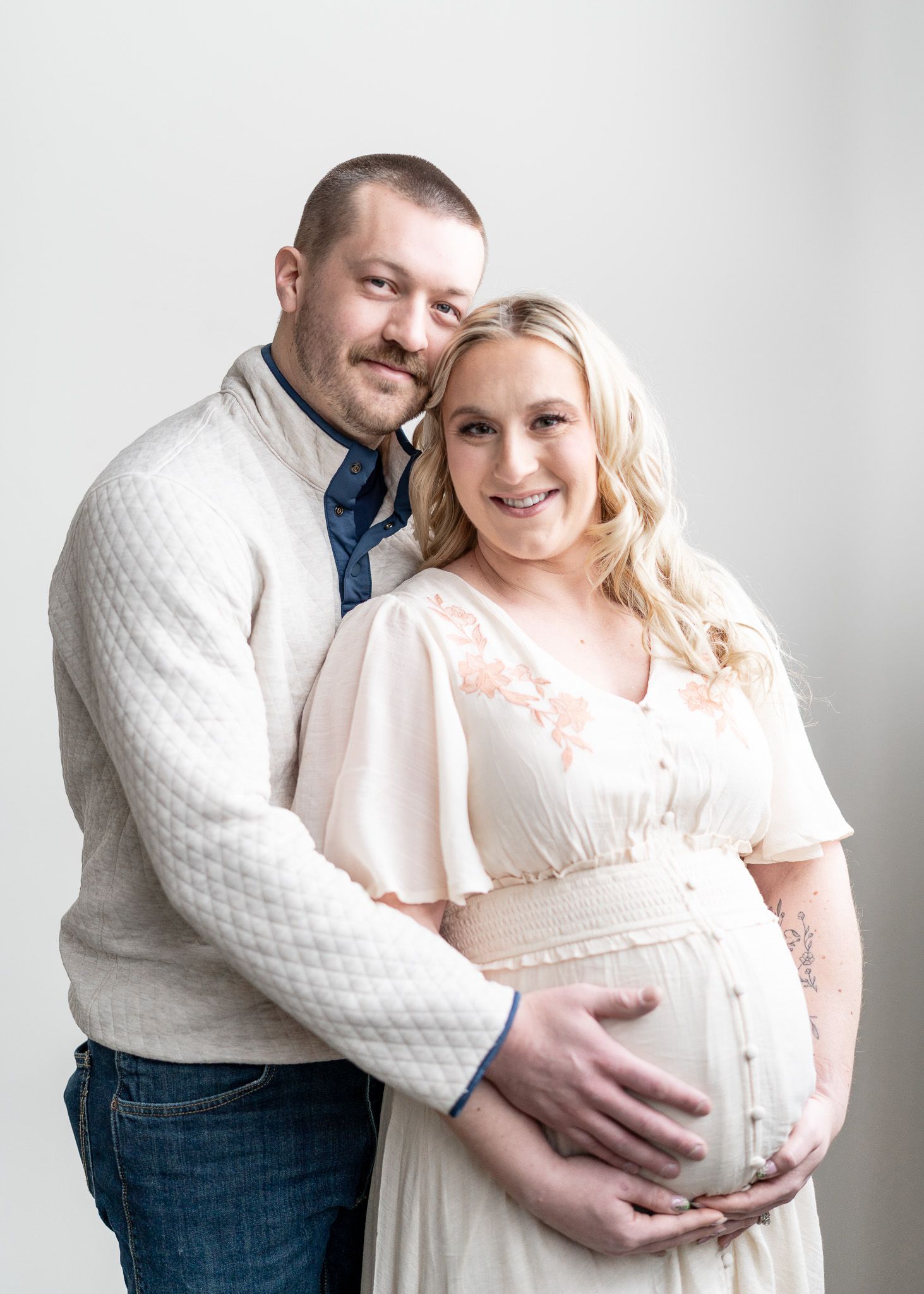 Intimate maternity portraits of couple in white and denim sharing tender moments against white backdrop.