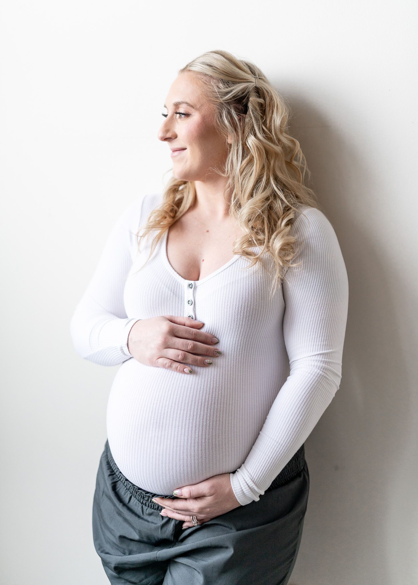 A pregnant woman in a white sweater poses against a white wall while holding her baby bump.