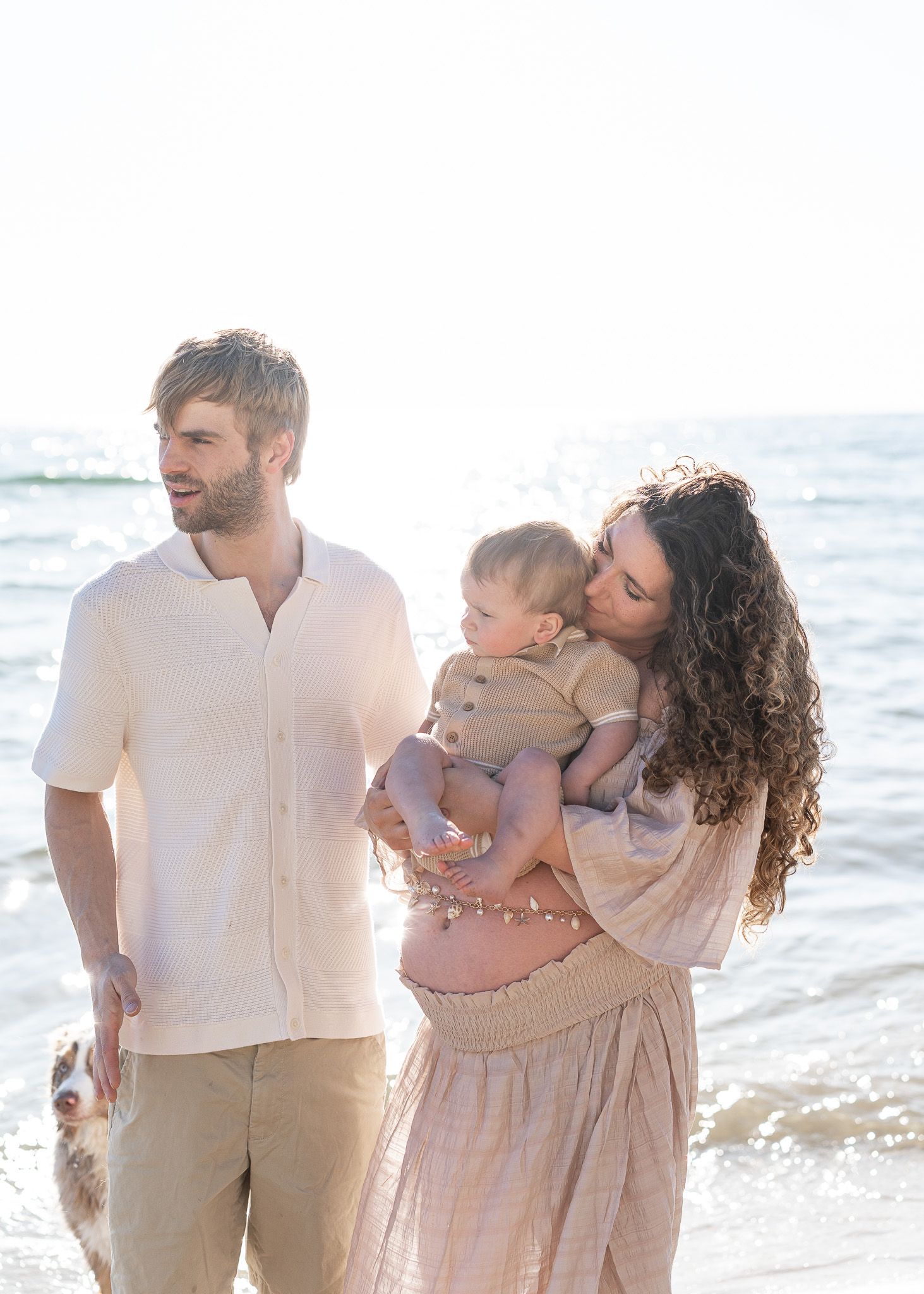Family in coordinated beige and white outfits shares tender moments together on a sunny beach at sunset.