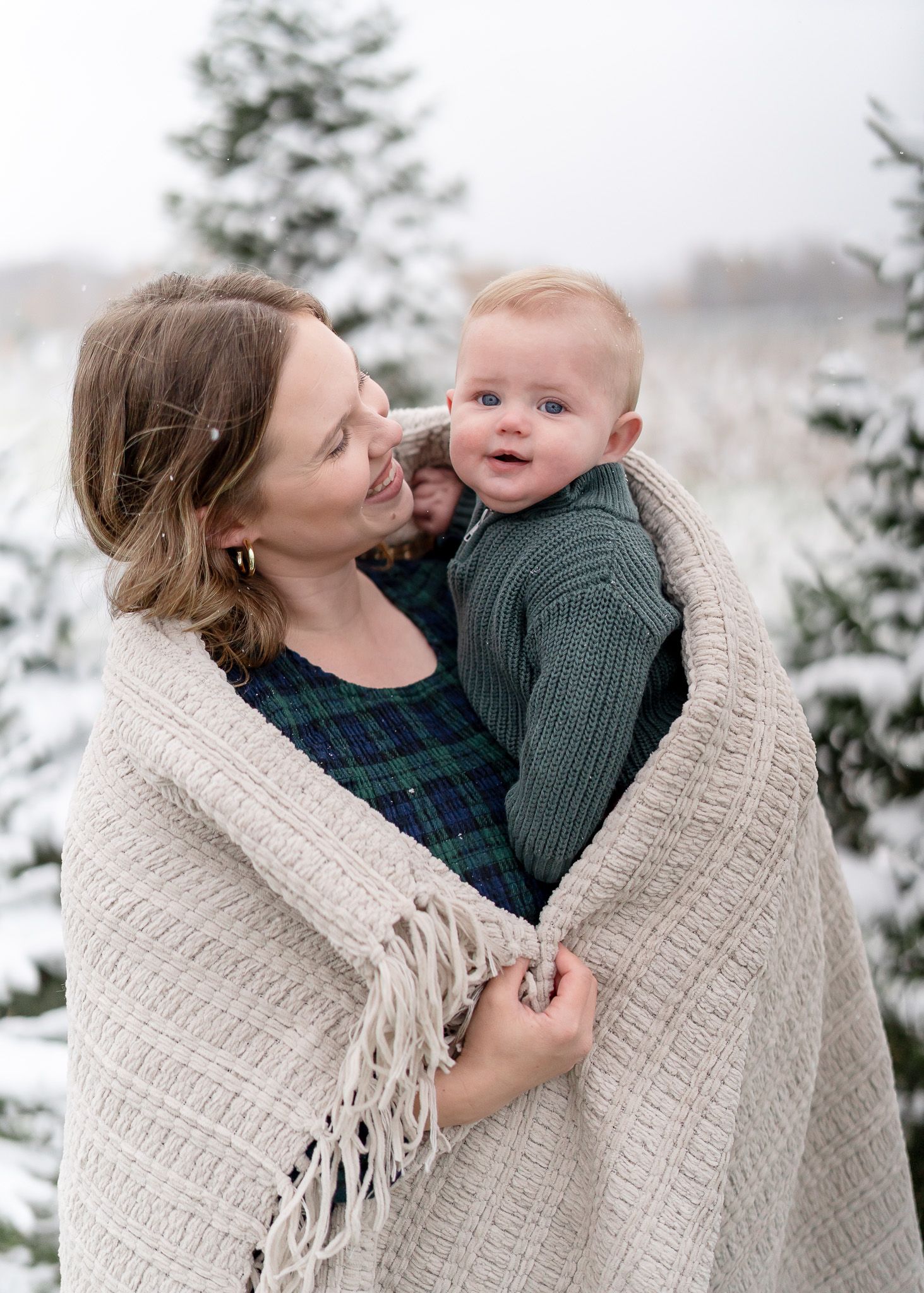 A tender winter moment captured outdoors with a cozy knit blanket wrapped around two people against a snowy evergreen backdrop.