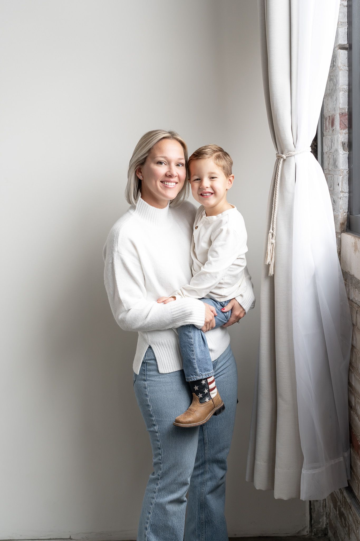 Parent and child in matching white sweaters share joyful moments against white curtains in a home photoshoot series.