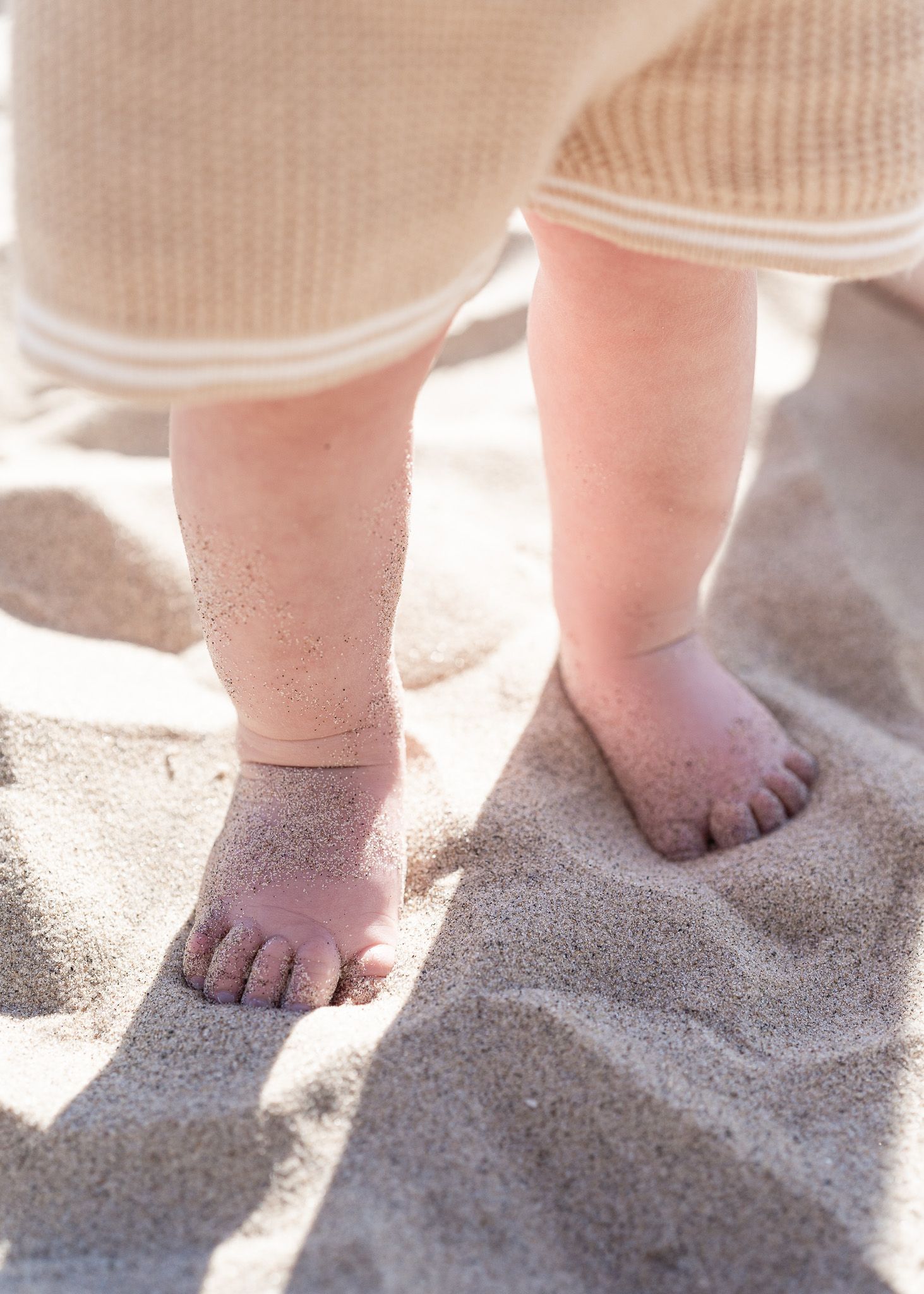 Small sandy feet of a baby standing on beach sand in soft sunlight with sand grains visible on the toes.
